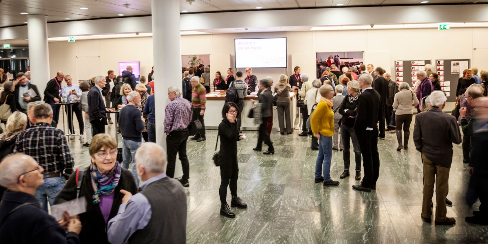 Crowd of people in a lobby of a venue, some standing at booths, with a screen in the background displaying 'Herzlich Willkommen'.