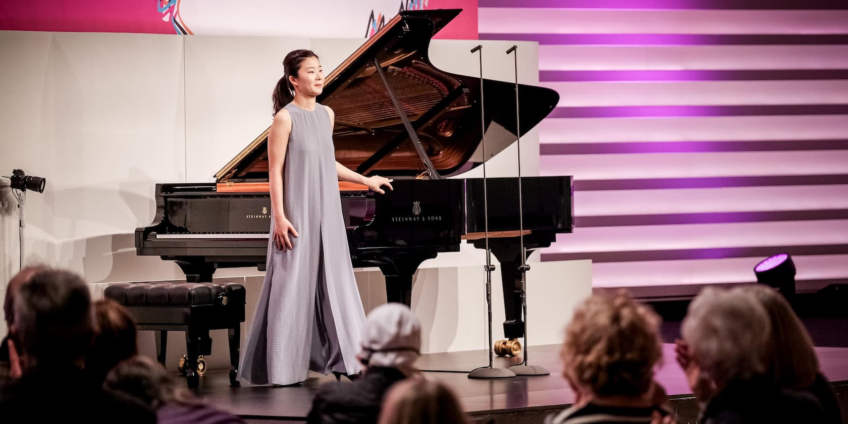 A woman stands next to a grand piano on a stage in front of an audience.