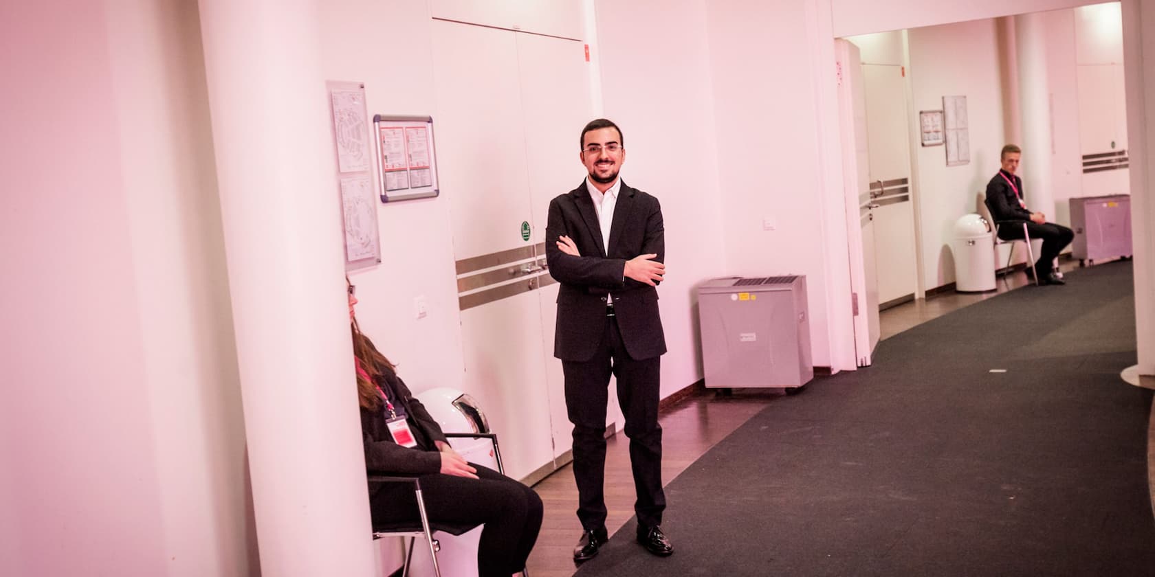 A man in a suit stands with crossed arms in a hallway, while a woman and another man sit on chairs.