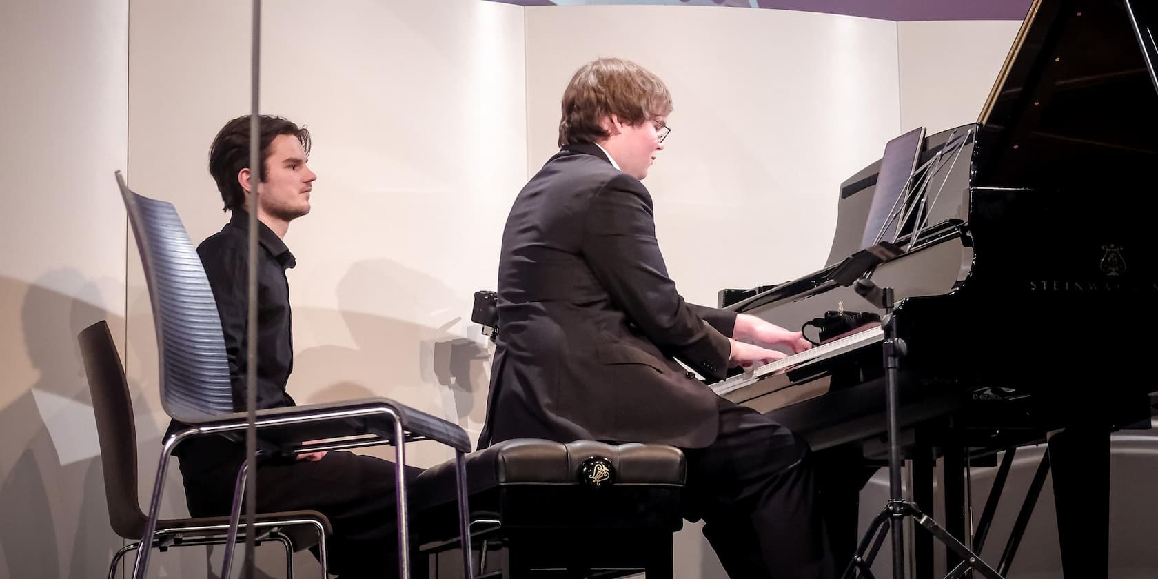 A pianist is playing the piano while a man sits next to him on a chair, watching.