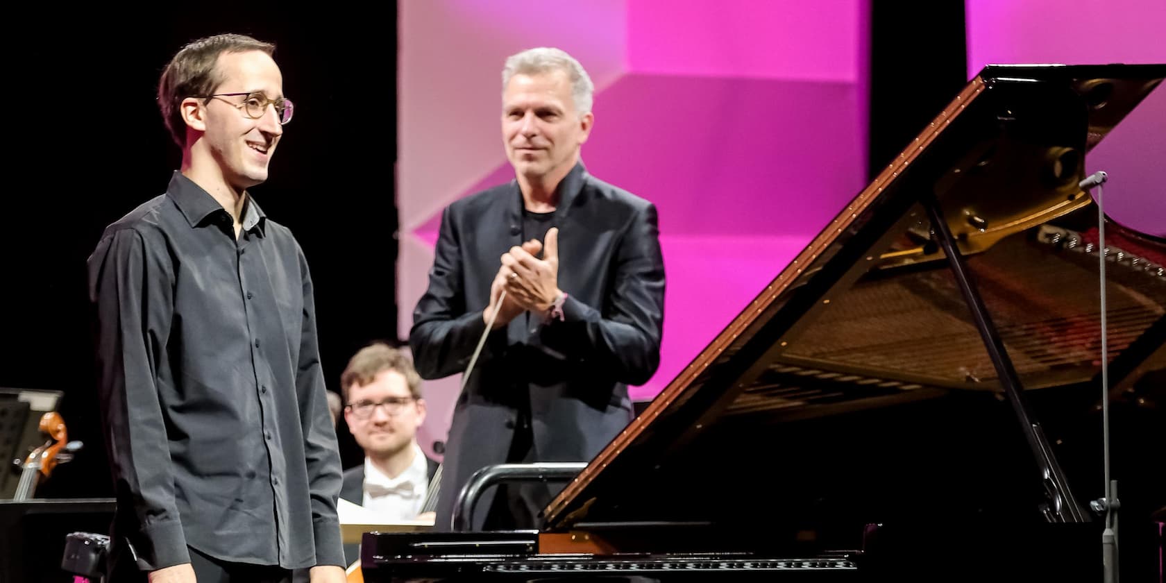 A musician stands next to a grand piano on a stage while a man in the background claps.