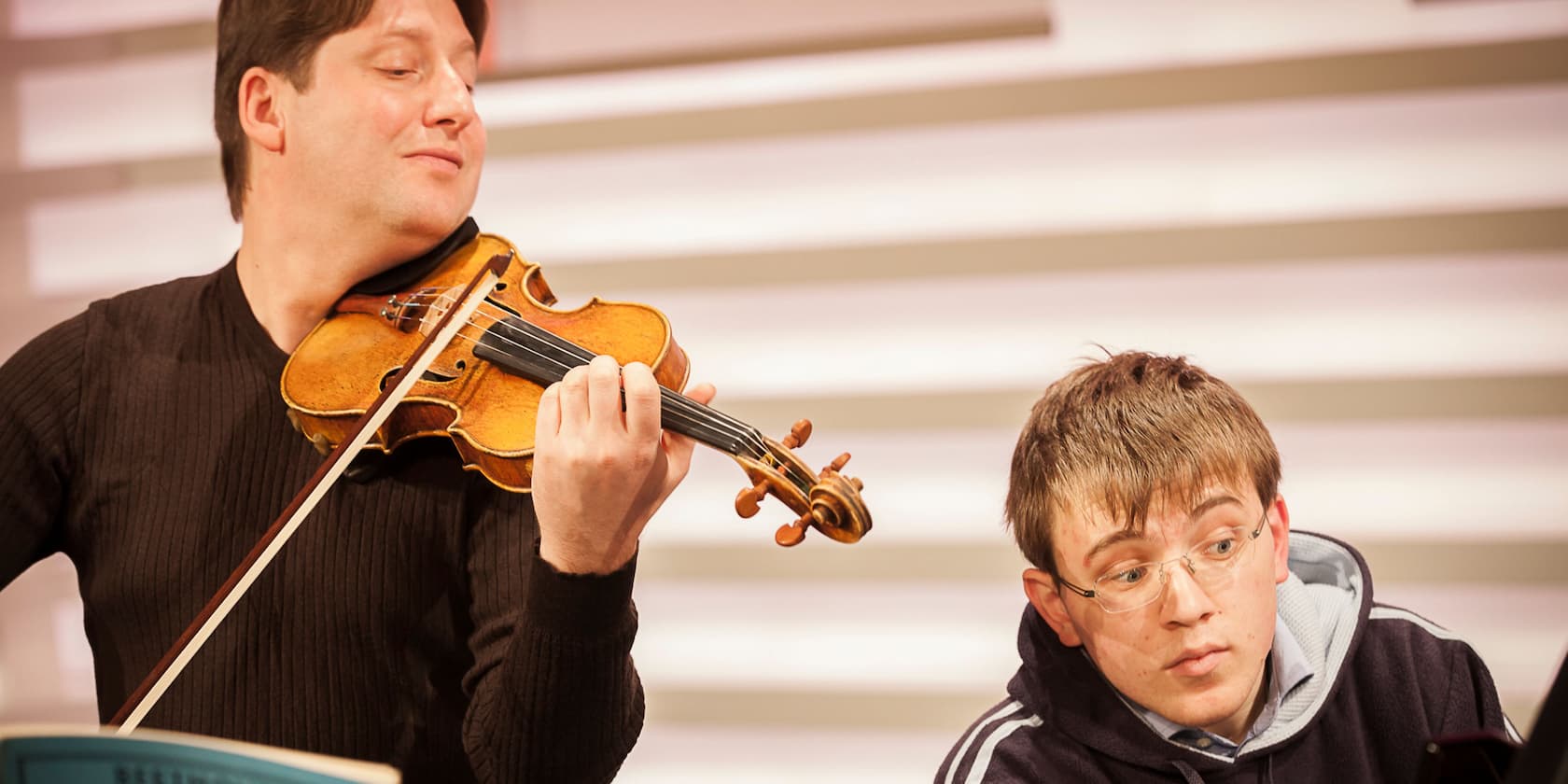 A man is playing the violin while another man with glasses watches intently.