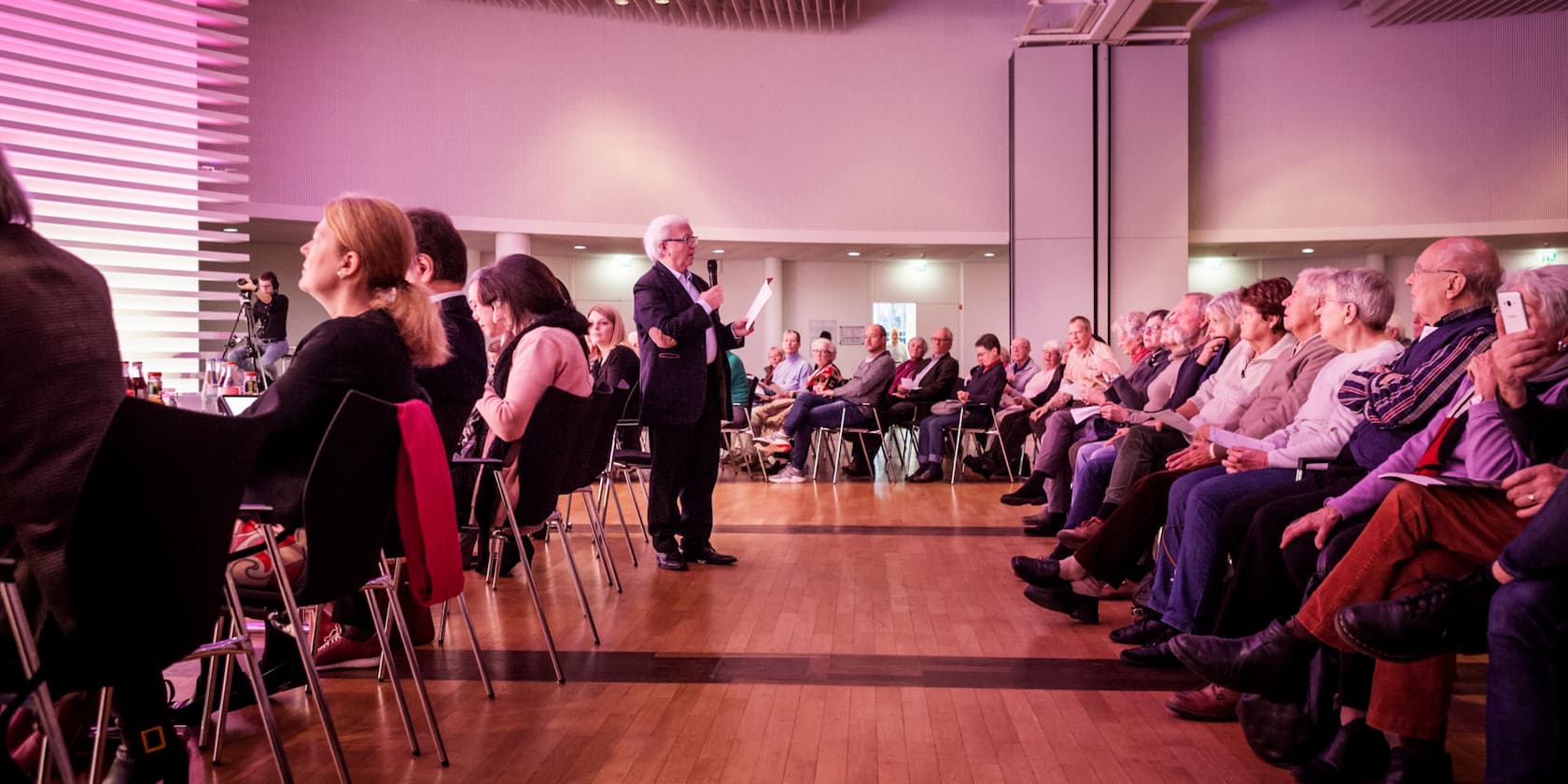 A man speaks into a microphone to a group of people seated in a conference room.