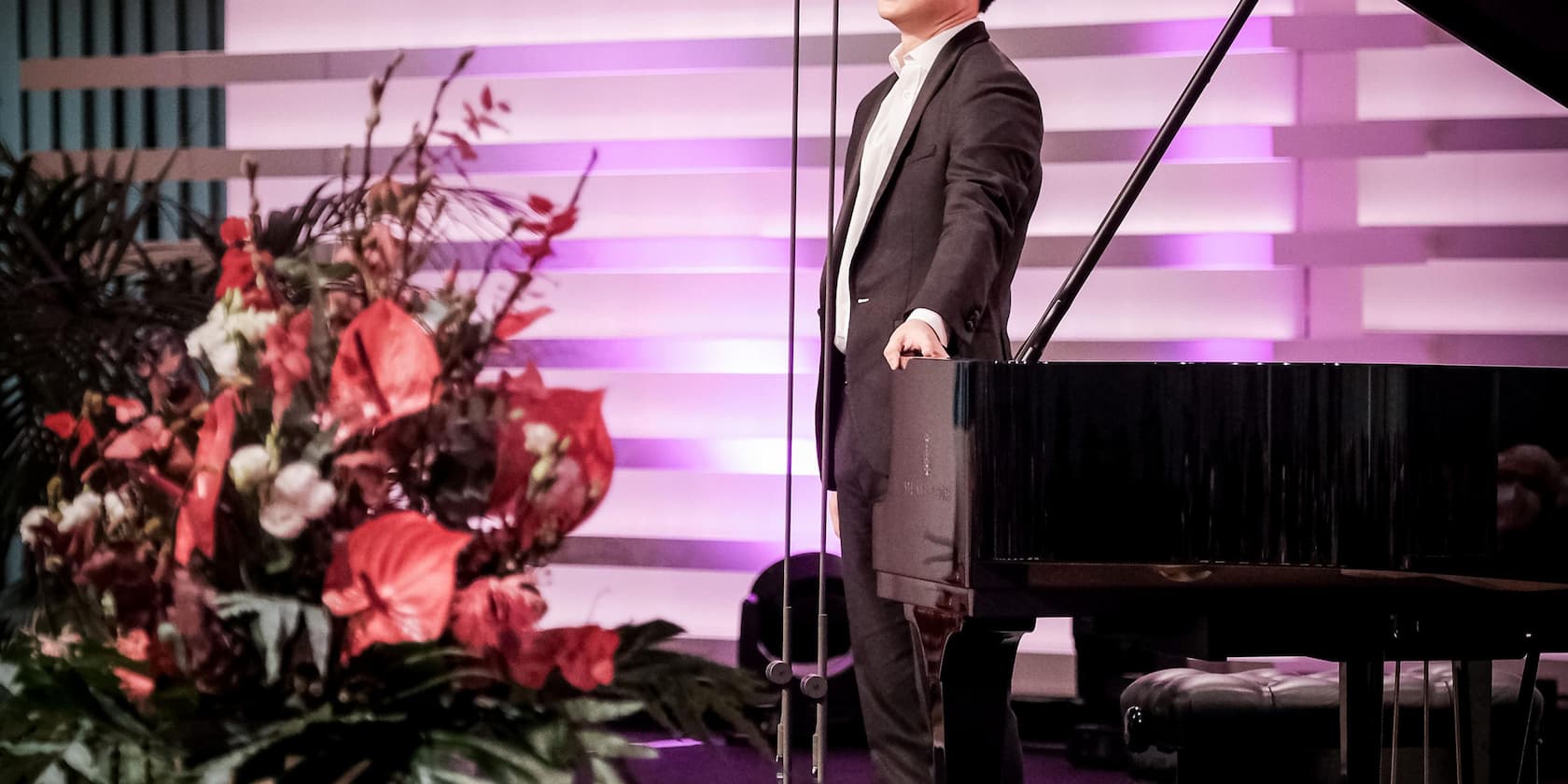 A musician in a suit stands next to a grand piano on a stage illuminated with pink lights. In the foreground on the left, there is a large flower arrangement.