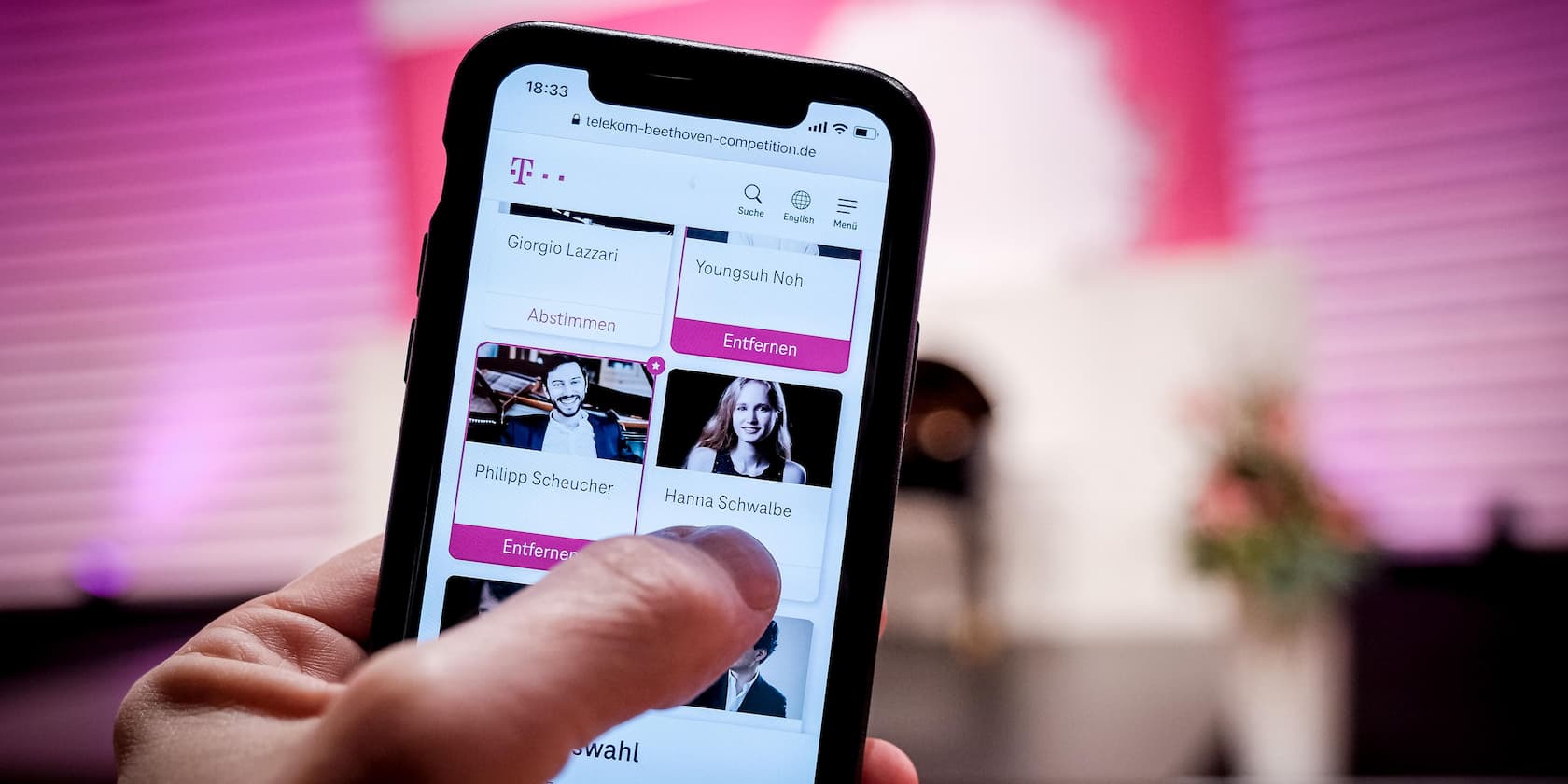 Eine Hand hält ein Smartphone, das die Webseite der Telekom Beethoven Competition zeigt, mit Optionen wie 'Abstimmen' und 'Entfernen' neben Bildern von Teilnehmern.