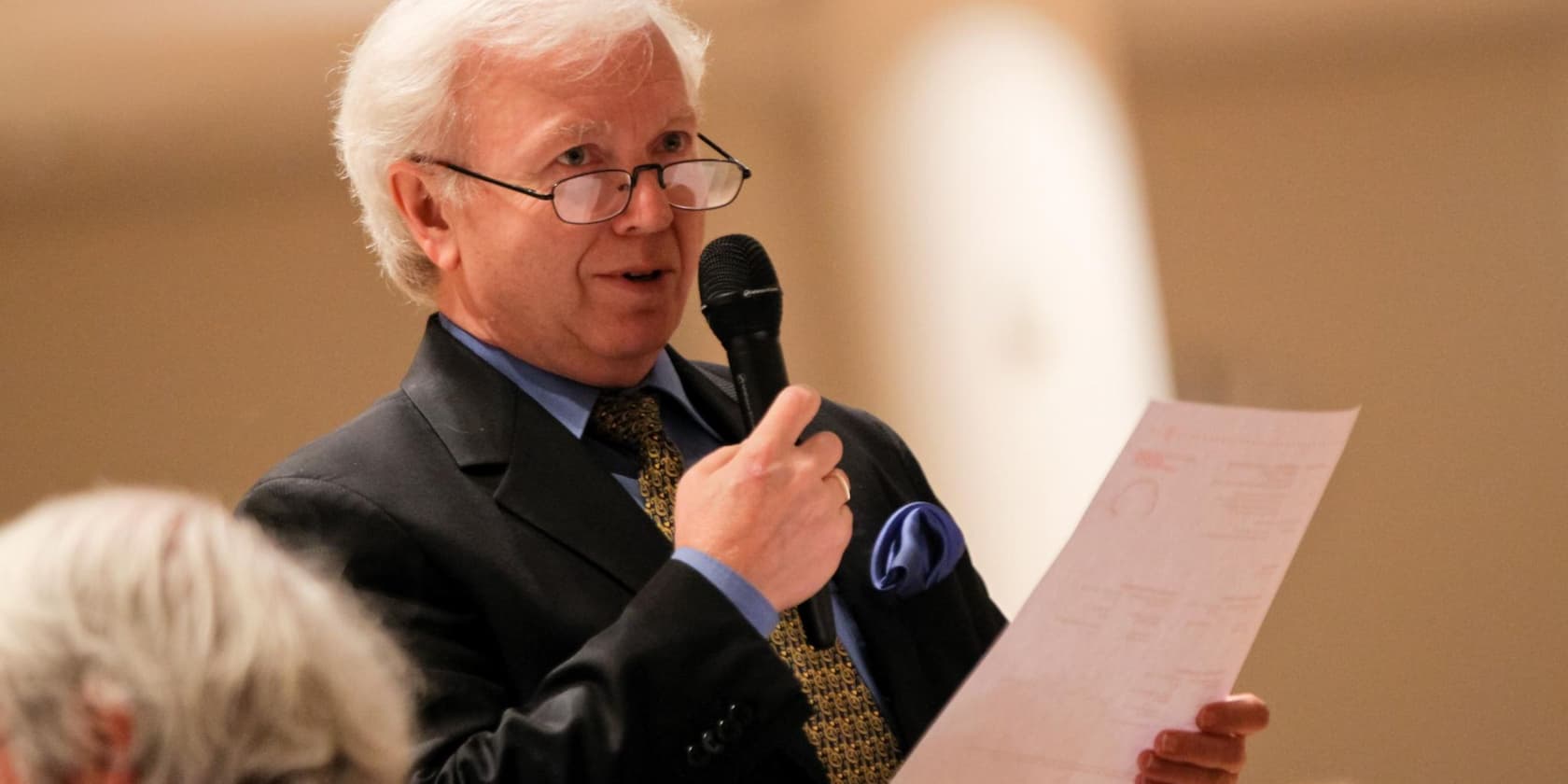 A man with glasses speaks into a microphone while holding a document in his hand.