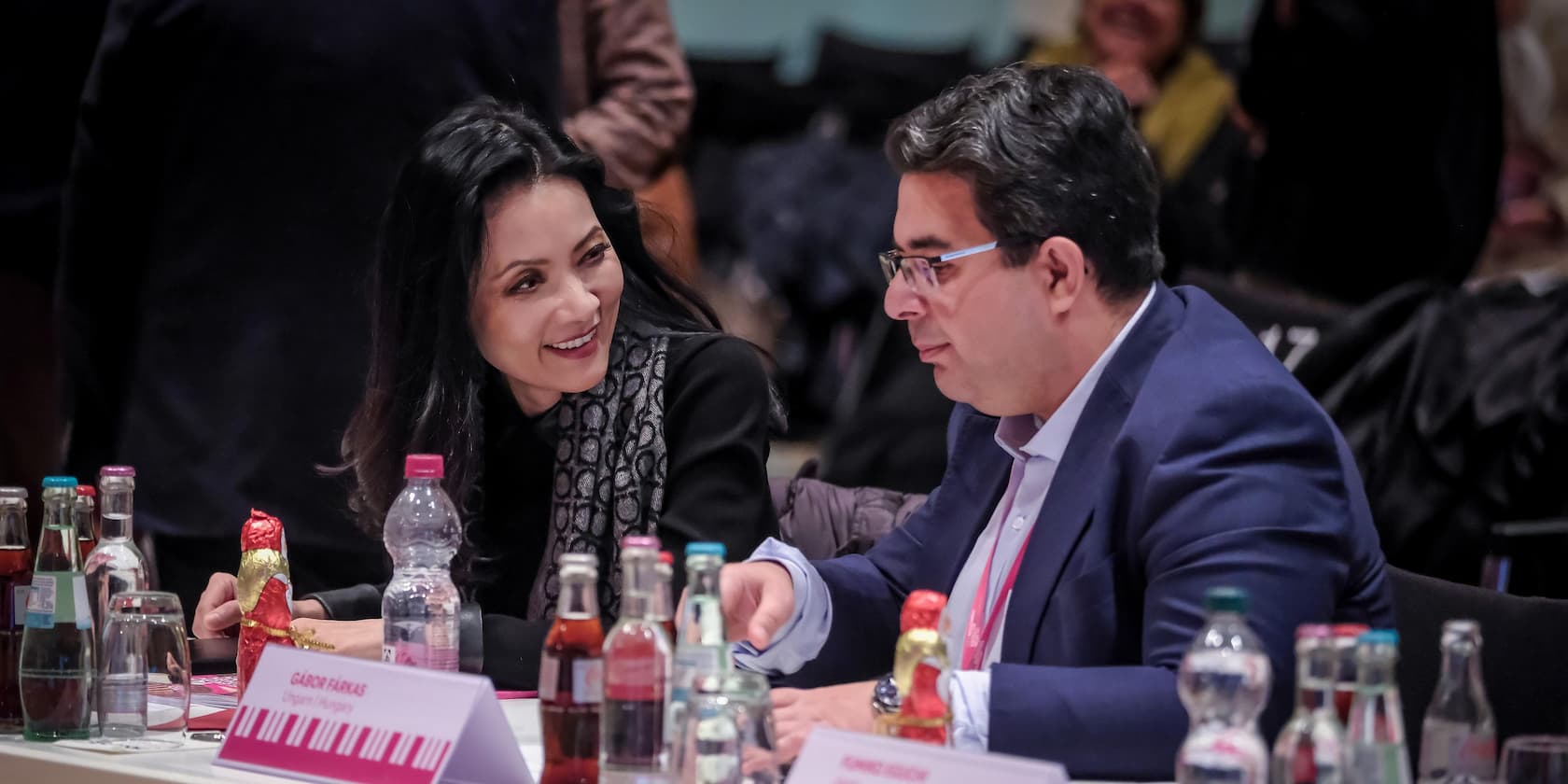 Two people are talking during a meeting. Several beverage bottles and a chocolate figure are on the table. A name tag with the inscription 'Gábor Farkas' is visible.