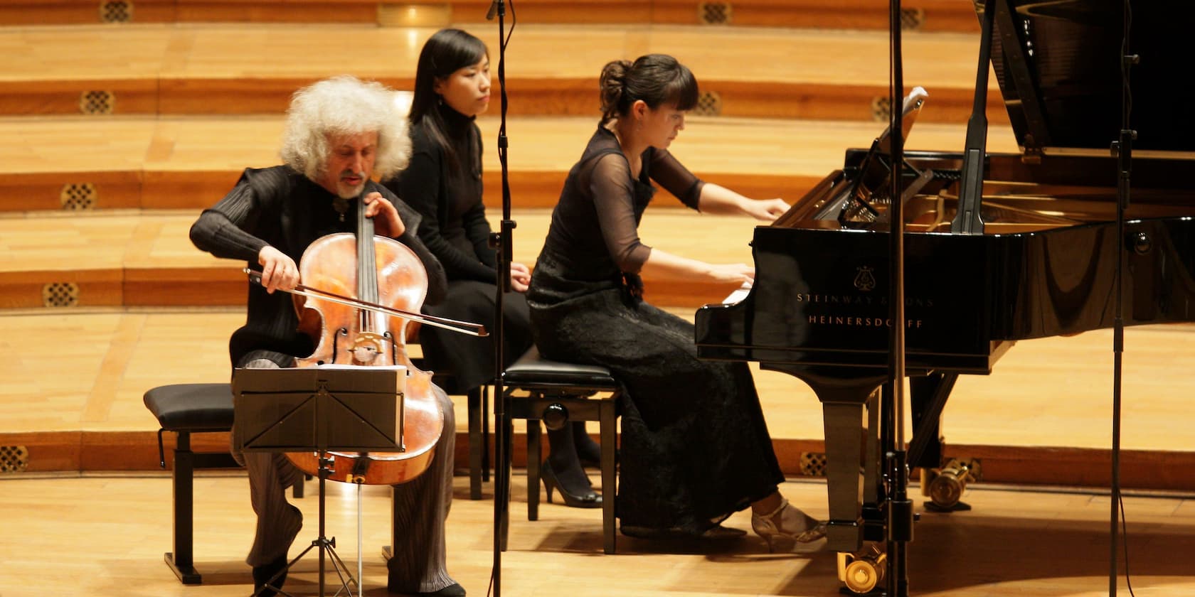 A cellist and a pianist perform on a stage, with another person sitting next to them.