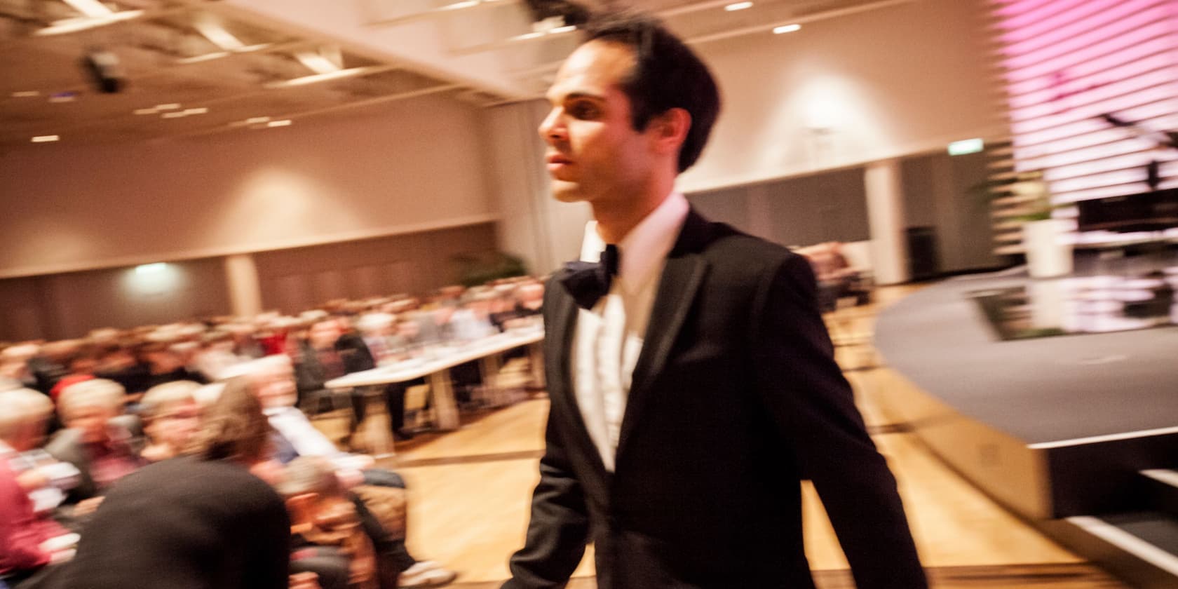 Blurred man in a suit walking through a room with a seated audience.