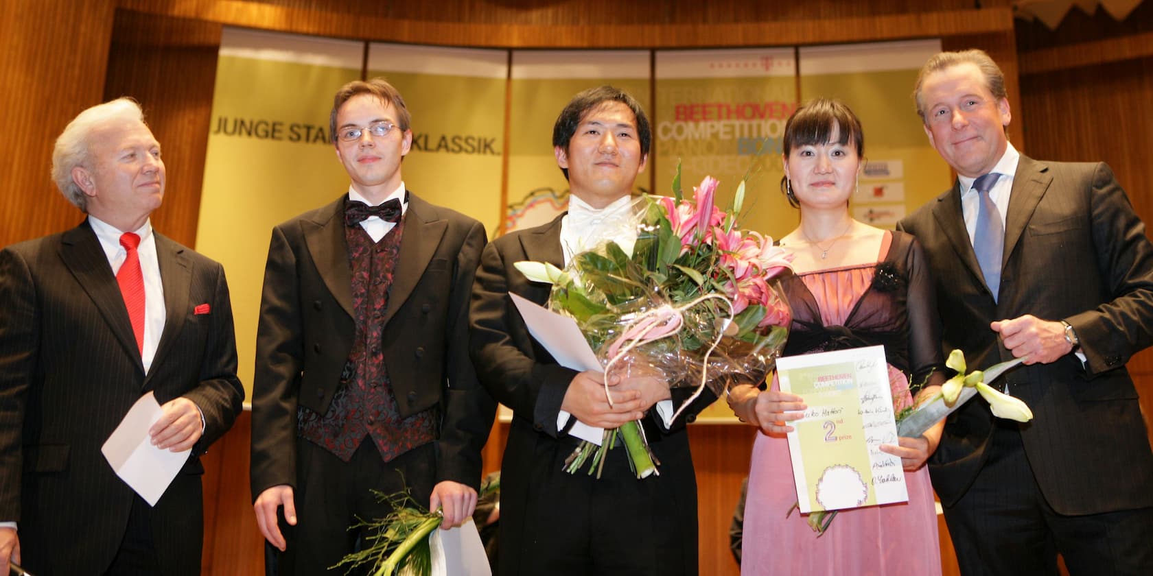 Fünf Menschen stehen auf einer Bühne bei einem Musikwettbewerb. Zwei der Teilnehmer halten Blumensträuße und Zertifikate. Im Hintergrund ist ein Schild mit der Aufschrift 'Beethoven Competition' zu sehen.