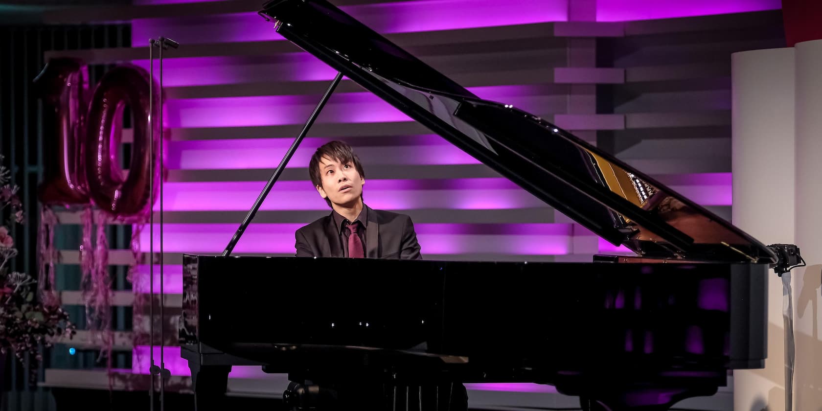 A man in a black suit plays the piano on a stage with purple lighting. Balloons forming the number 10 are visible in the background.