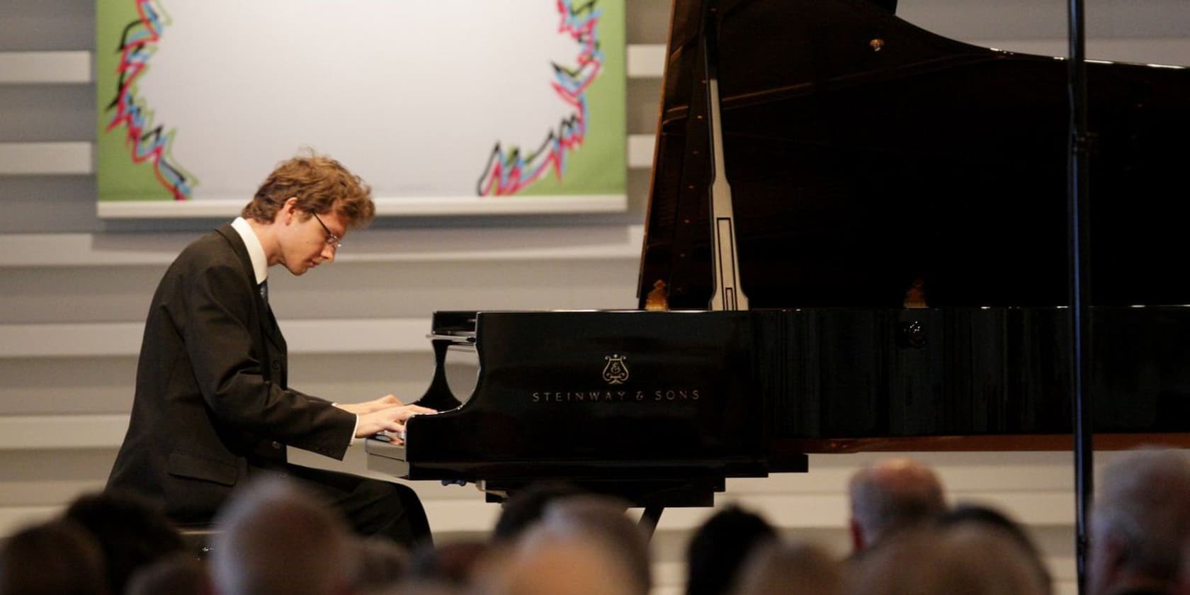 A man in a suit plays a piano at a concert, seated in front of an audience.