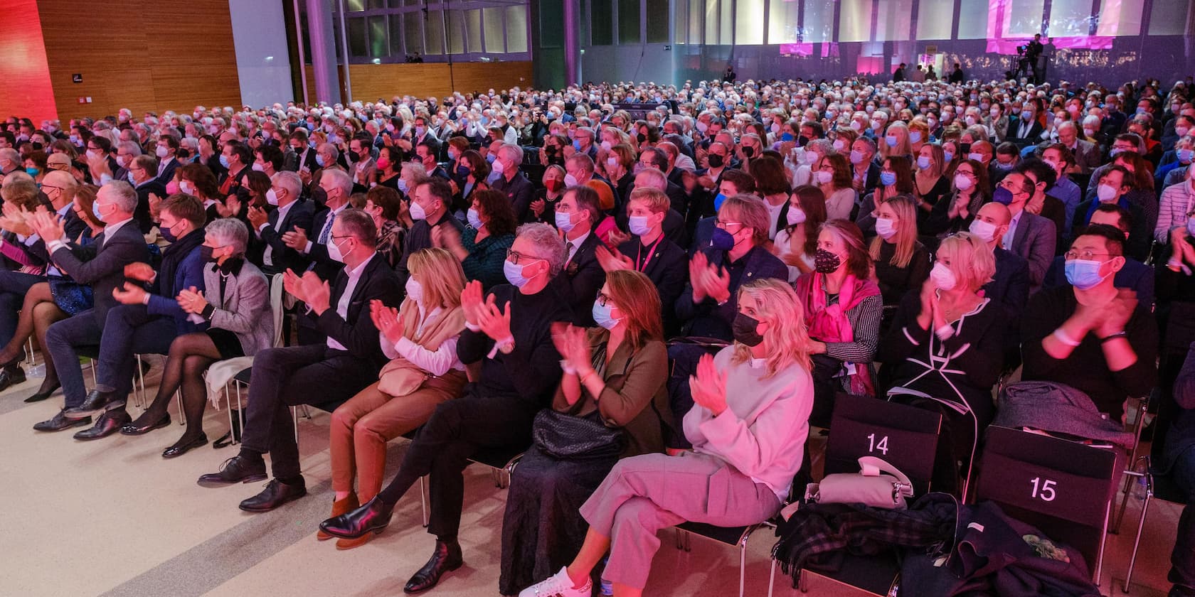 A large crowd sits in an auditorium clapping, many wearing face masks.