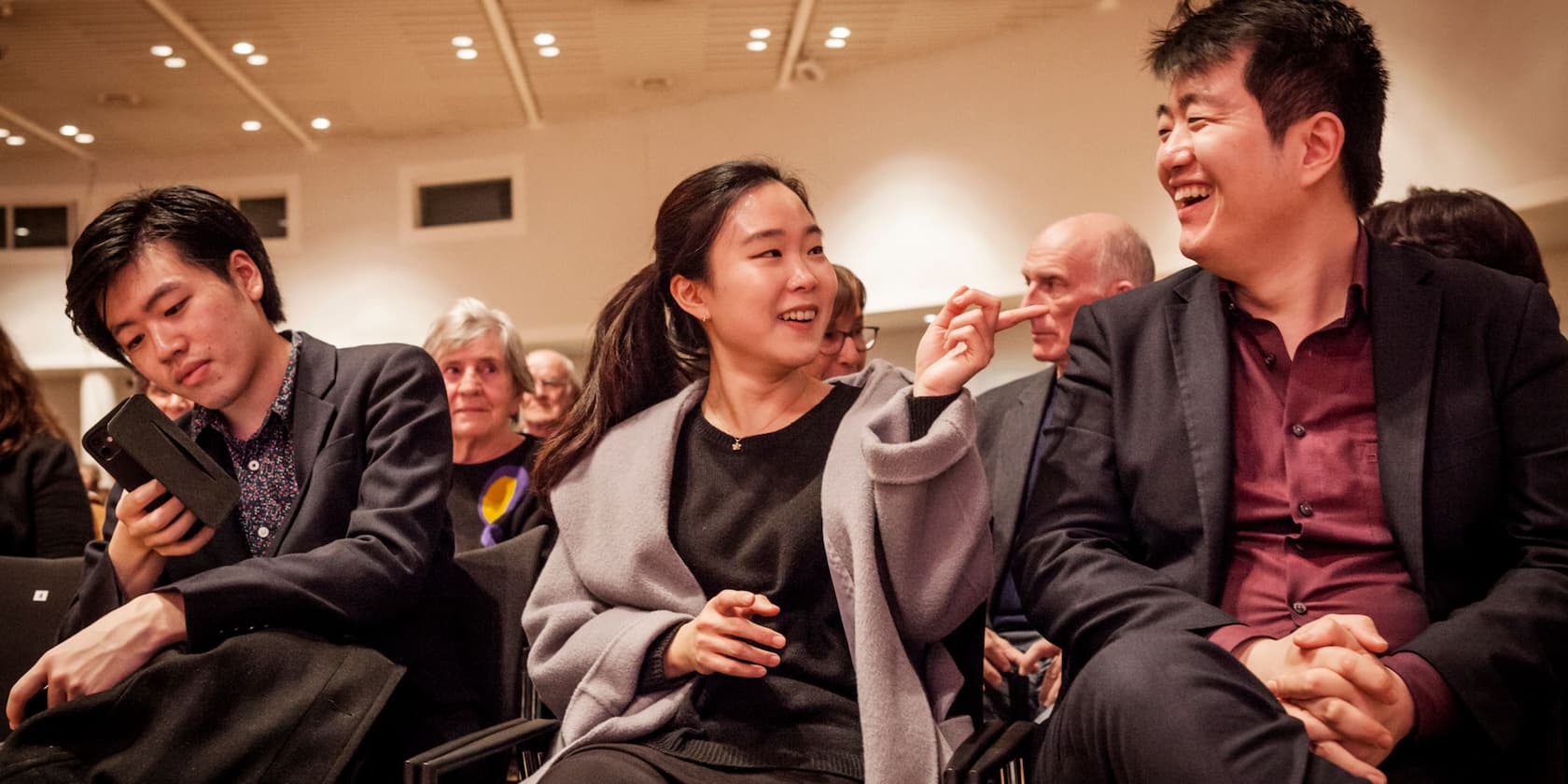 A group of people sitting in a room. A woman is pointing at something and smiling with a person next to her. A man next to them is looking at his phone.