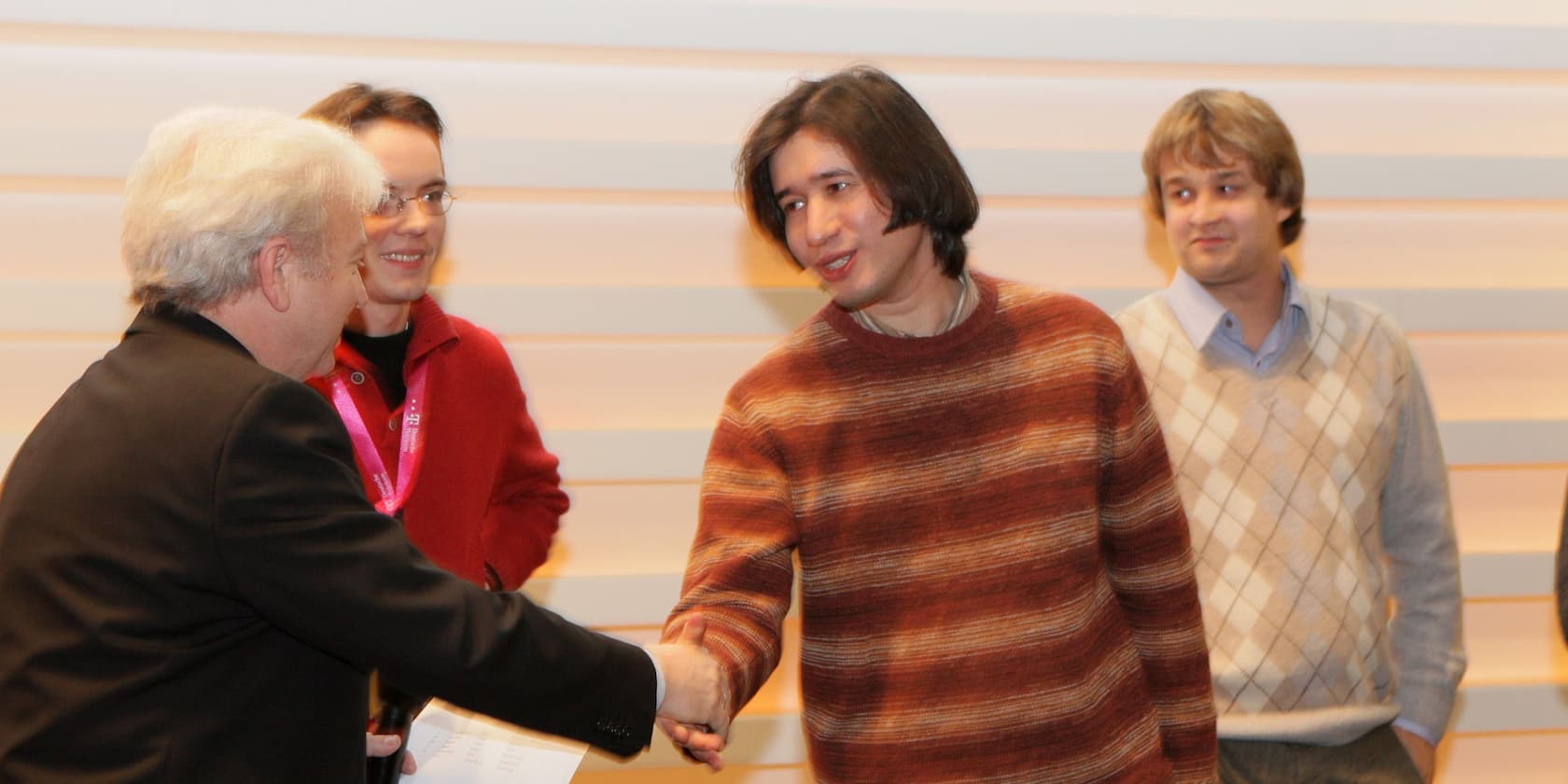 A person in a suit shakes hands with a man in a brown sweater, while two other people watch.