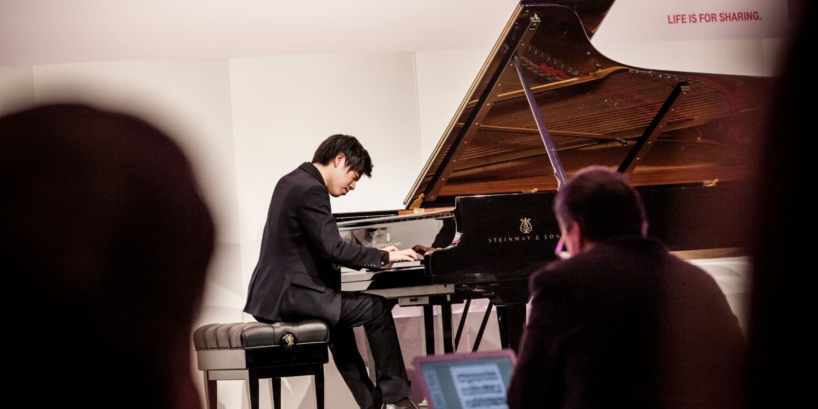 A man playing piano on stage, with the inscription 'LIFE IS FOR SHARING.' in the background.