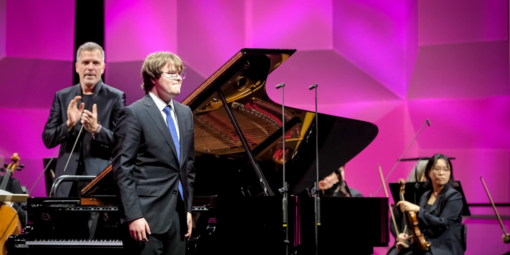 A pianist in a suit bows on a stage while the conductor applauds and a violinist sits in the background. The background is lit in pink.