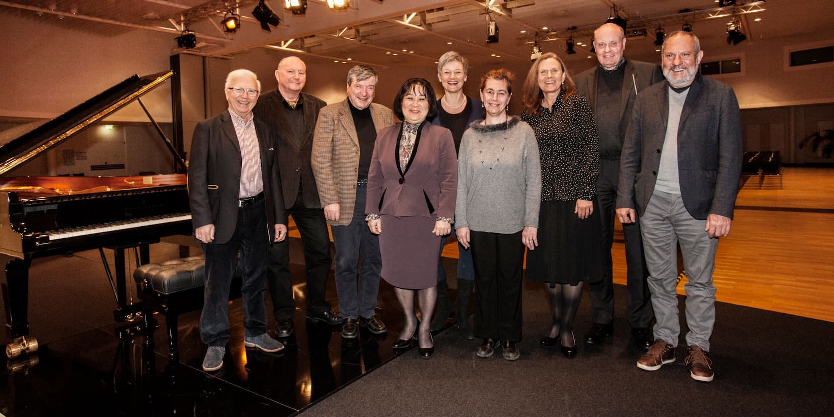 A group of nine people standing in front of a grand piano in a well-lit room.