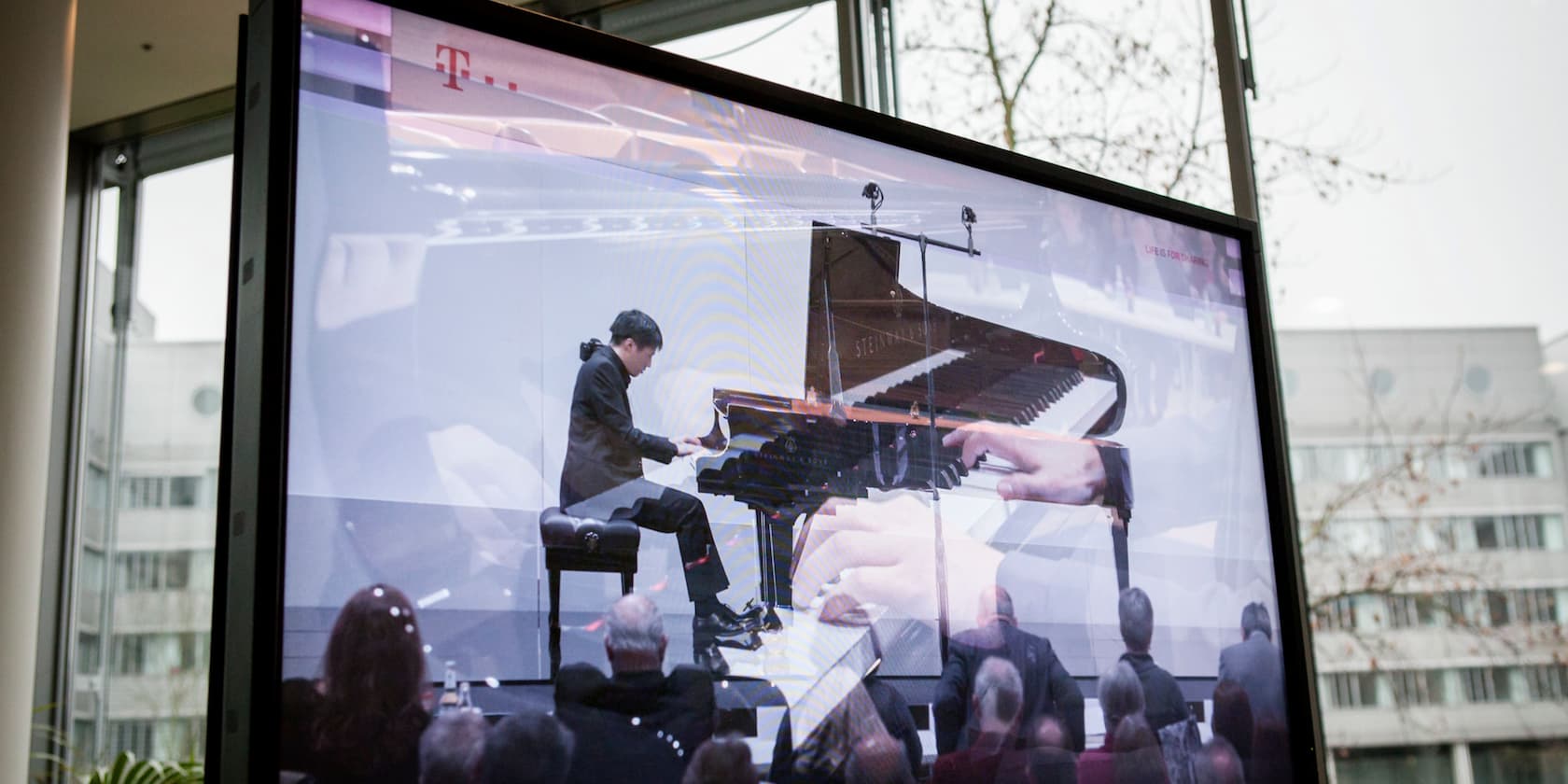 Ein Pianist spielt vor Publikum auf einem Flügel. Das Bild wird auf einem großen Bildschirm gezeigt.