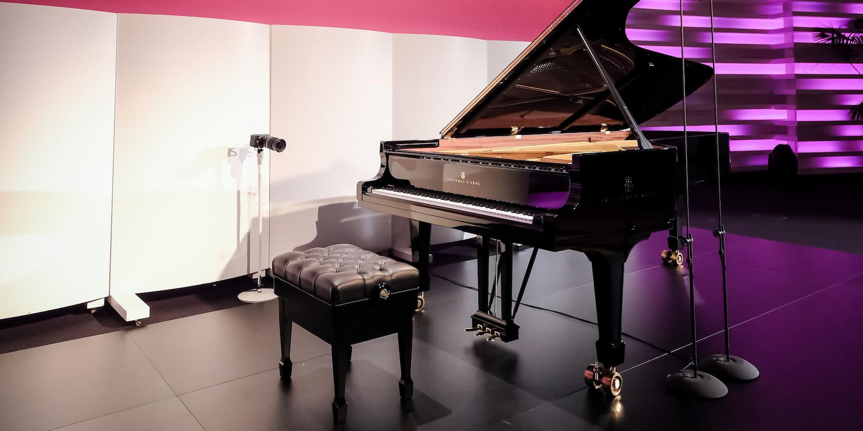 A black grand piano and a black piano bench on a stage with pink and white background lighting.