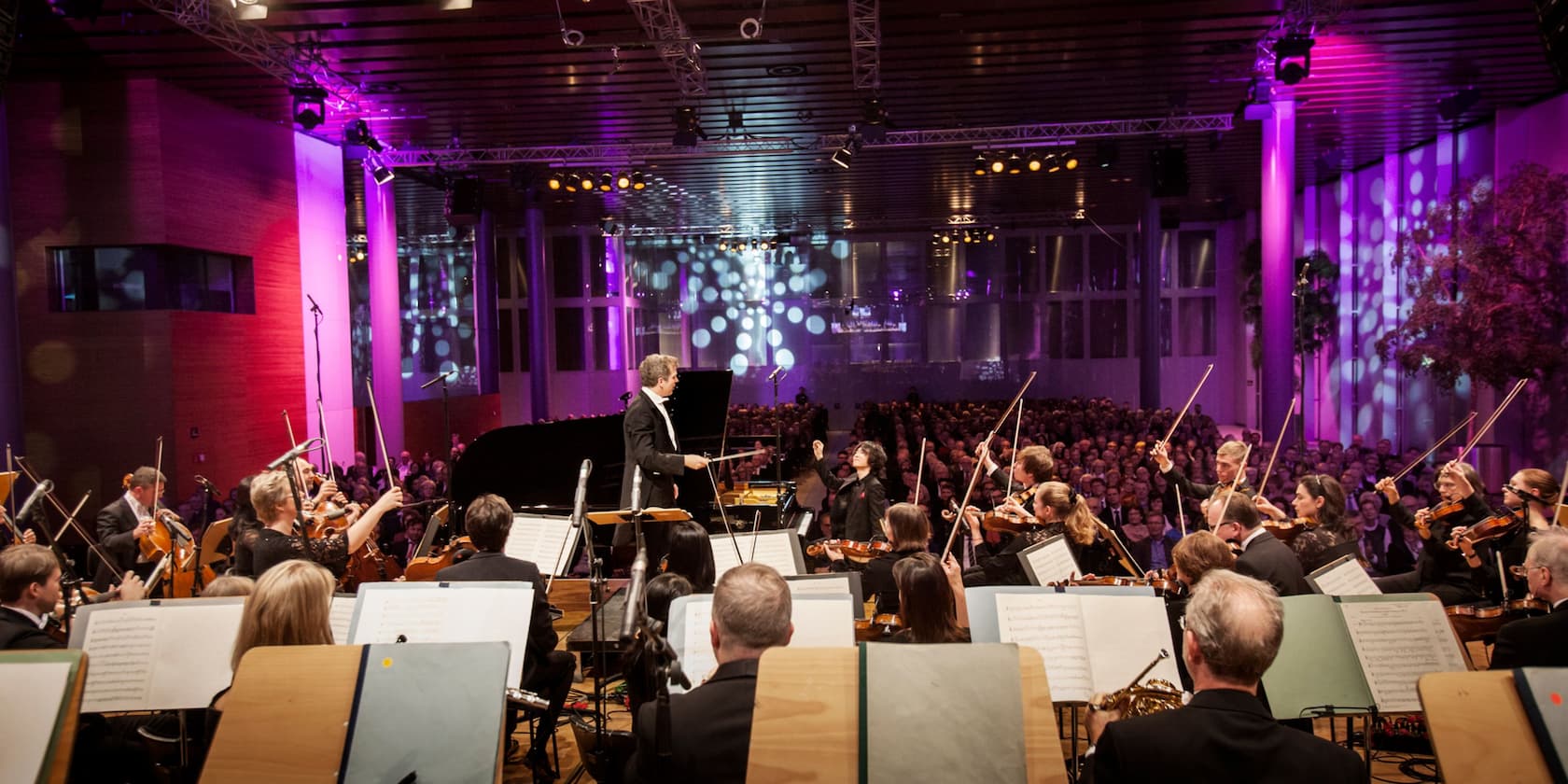 Ein Orchester tritt in einer großen, modernen Konzerthalle vor einem Publikum auf. Bunte Lichter beleuchten die Bühne.