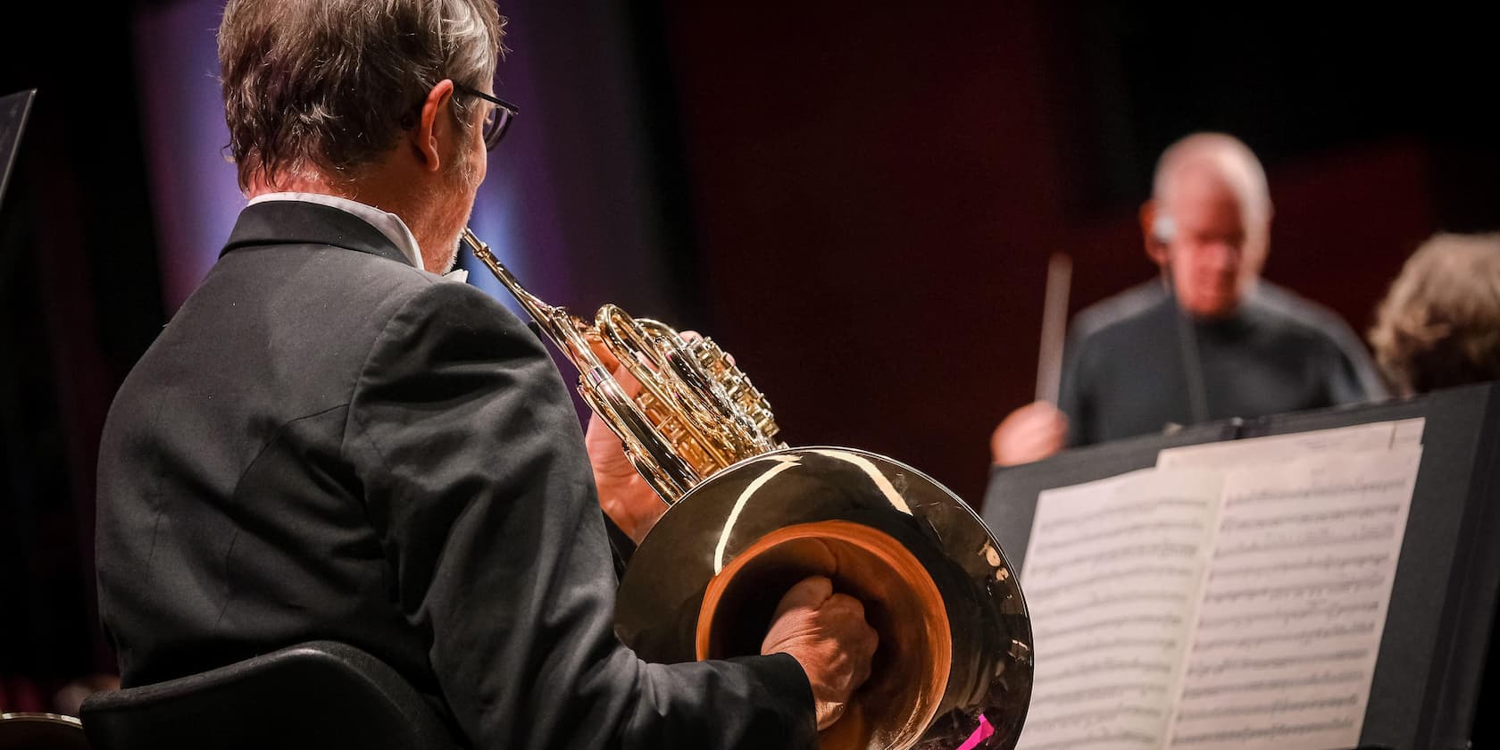 Musician playing a horn on stage, music stand in the foreground.