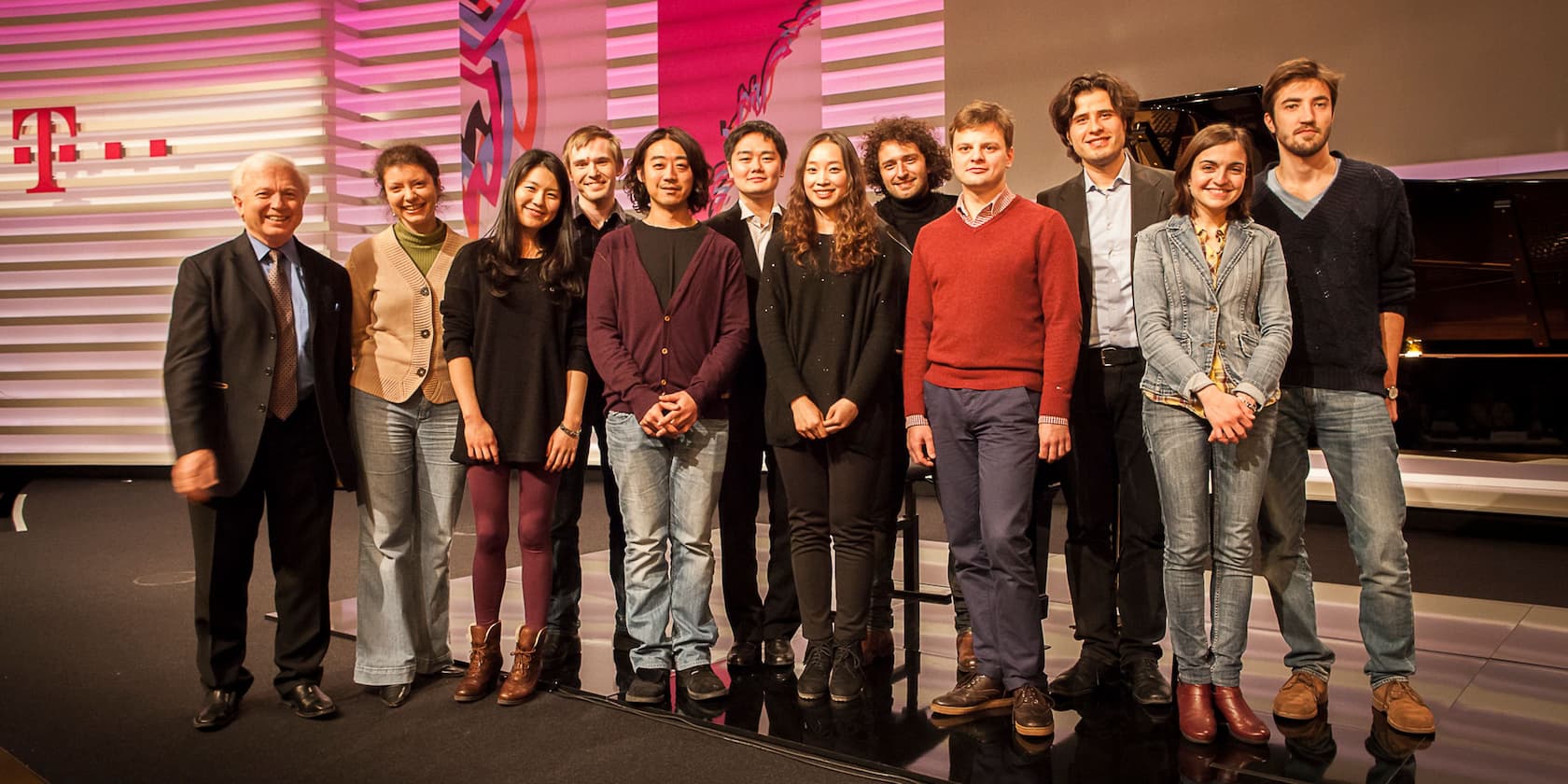 A group of twelve people standing on a stage in front of a background with a large T symbol and colorful patterns.