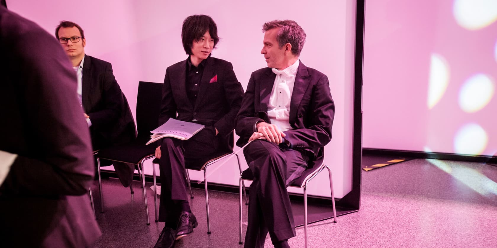 Three men in suits sit next to each other and converse in a room with pink lighting.