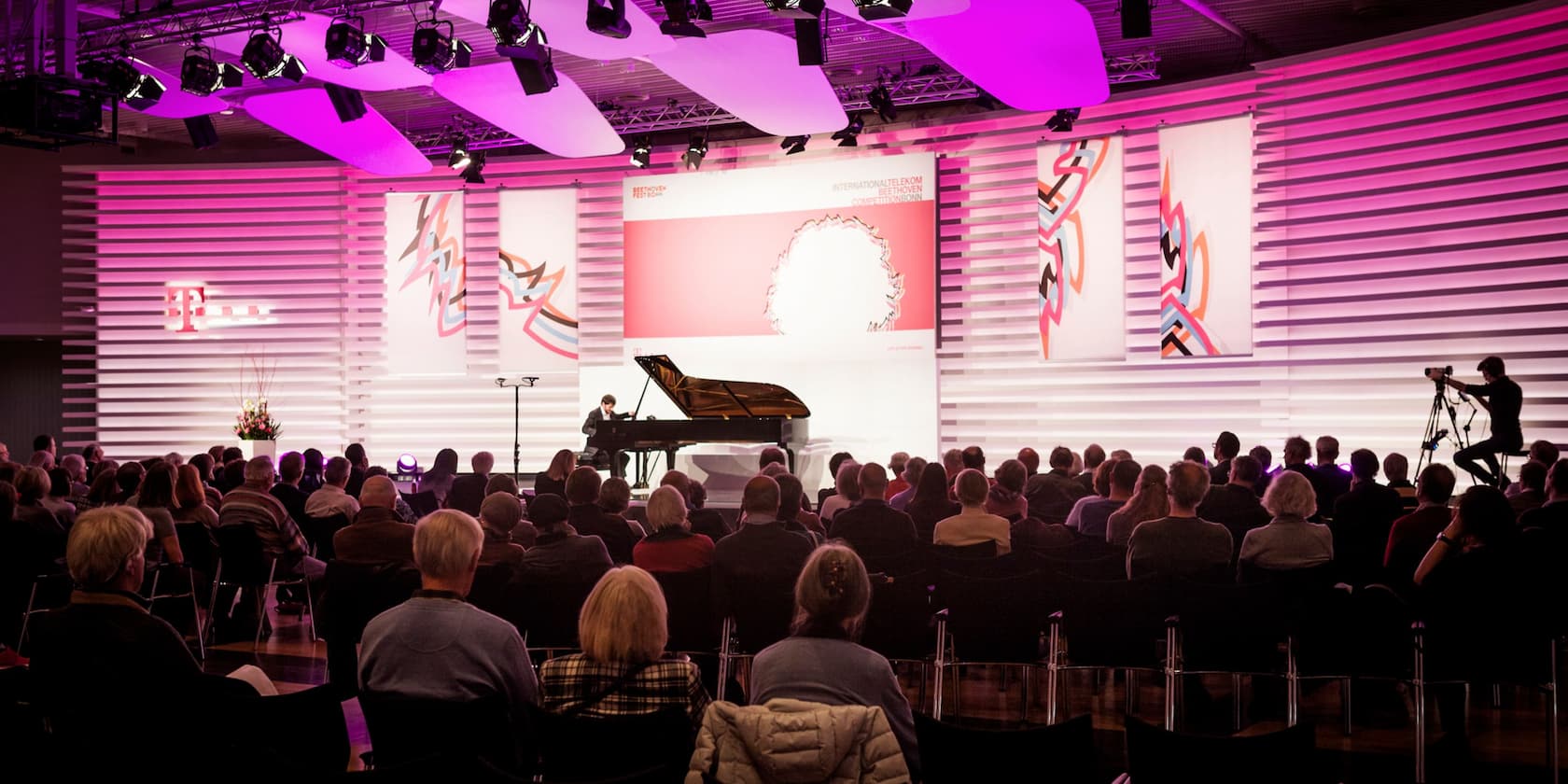 A concert with a pianist on stage, illuminated by pink lights, in front of an audience. A cameraman is filming the event.