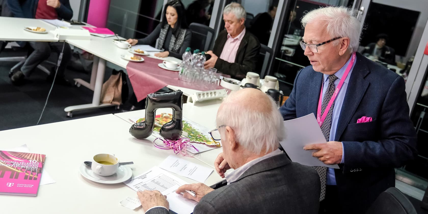 Four people in a meeting around a conference table. A man is standing and holding documents. On the table are snacks, drinks, and notes.