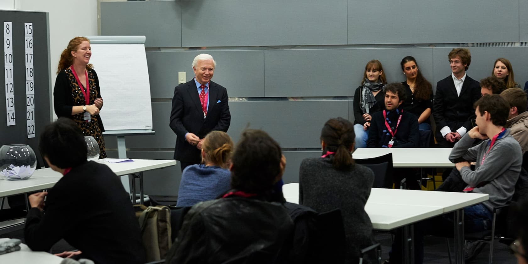 Eine Gruppe von Menschen sitzt und steht in einem Seminarraum. Zwei Personen, ein Mann und eine Frau, sprechen vor einem Schreibtisch mit einem Flipchart im Hintergrund.