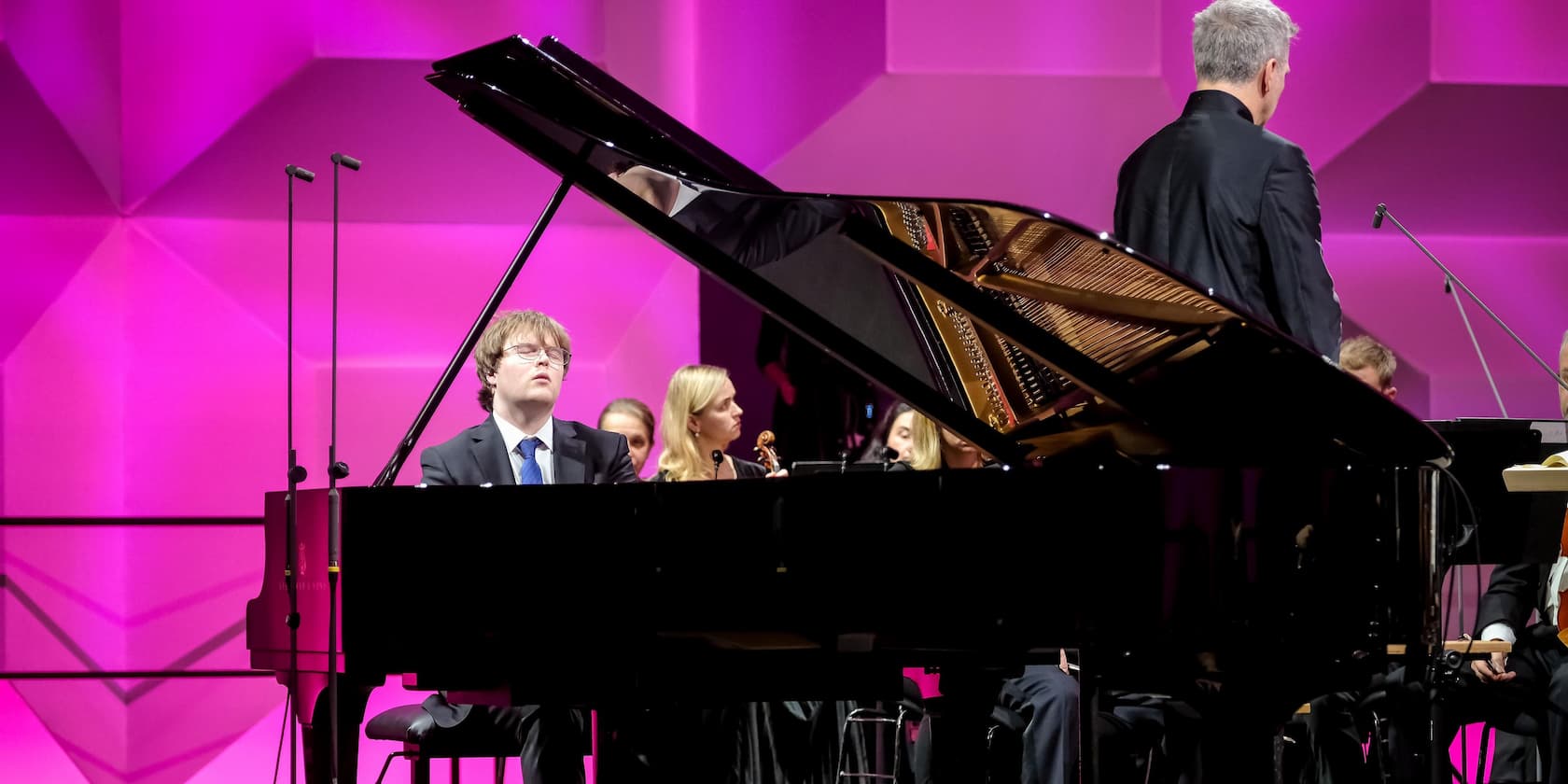 Pianist playing a grand piano with an orchestra in the background, stage lit in pink.