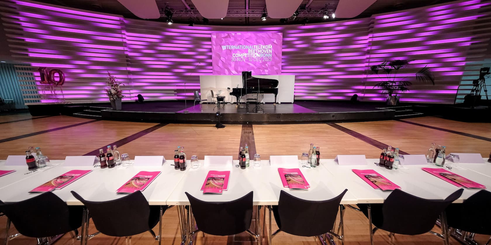 Konferenzraum mit Bühne und Klavier, lila Beleuchtung und Banner mit Text 'International Telekom Beethoven Competition Bonn 30 Nov - 10 Dec 2023'.