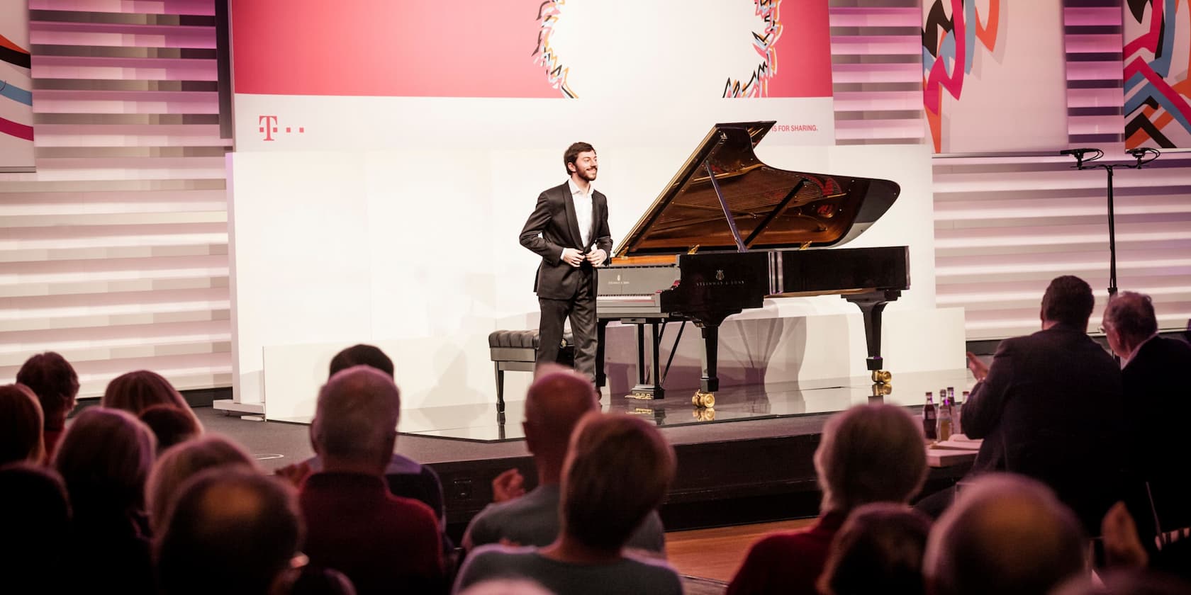 A pianist stands beside a grand piano on a stage, applauding the audience.