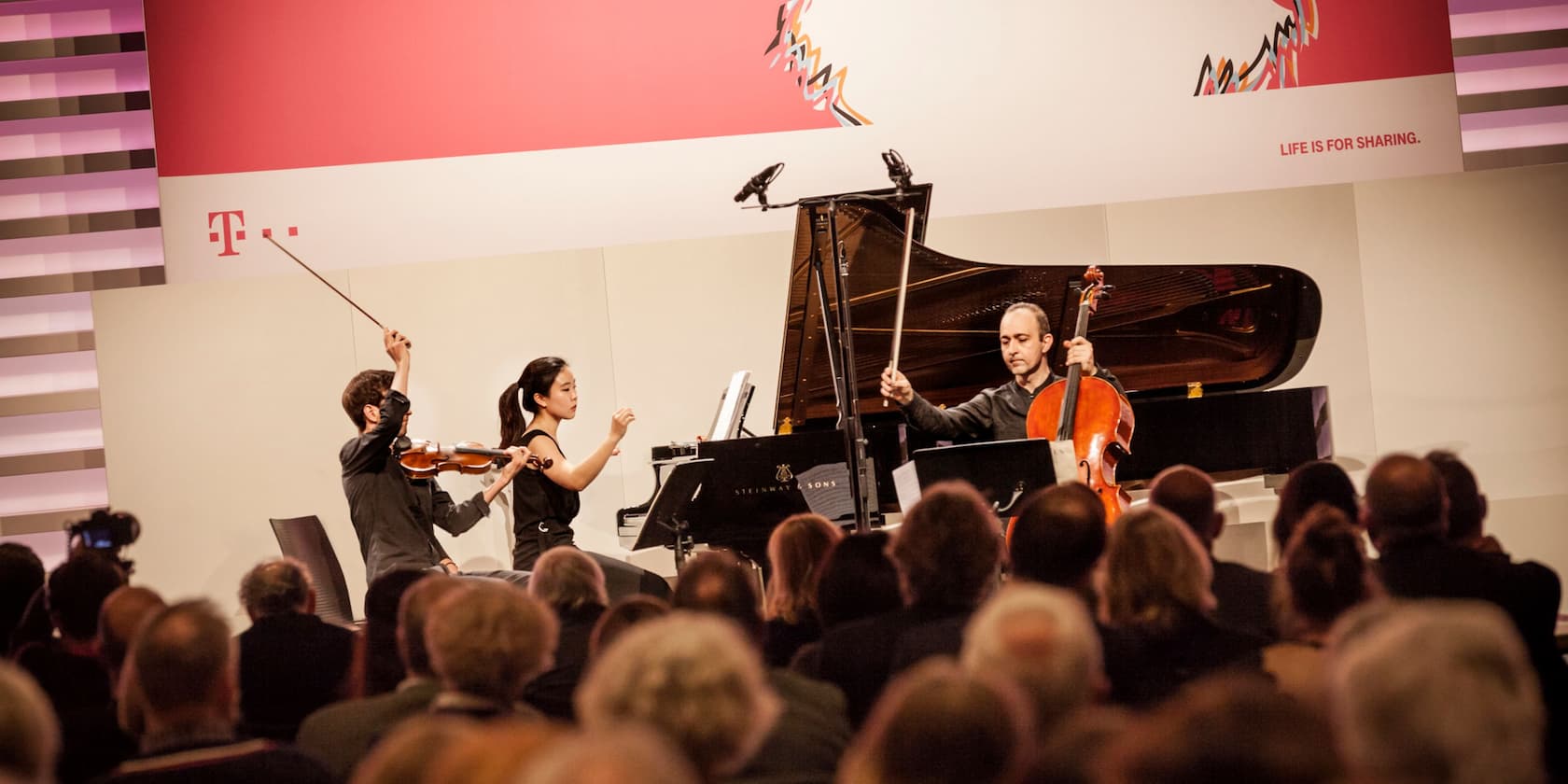 Musicians perform on stage in front of an audience, with the Deutsche Telekom logo visible in the background.