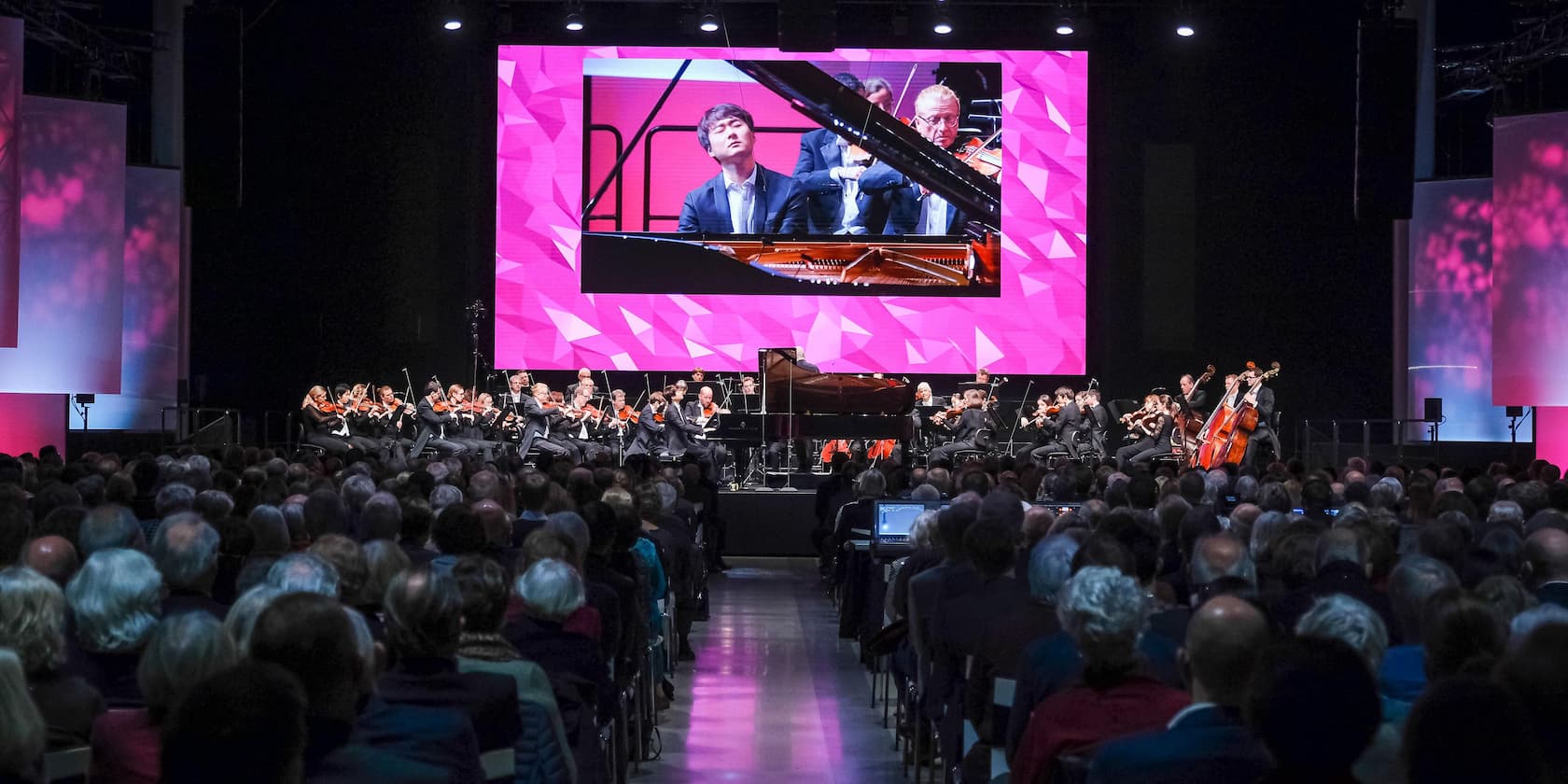 An orchestra performs on a stage before a large audience. A close-up of the pianist is displayed on a large screen above the stage.