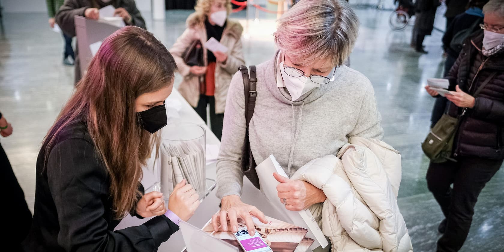 Two women wearing face masks are looking at a document or magazine together at a table during an event.