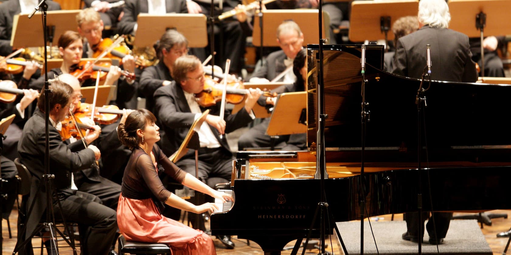 Eine Pianistin spielt in einem Orchester, umgeben von Violinisten und anderen Musikern auf einer Bühne.