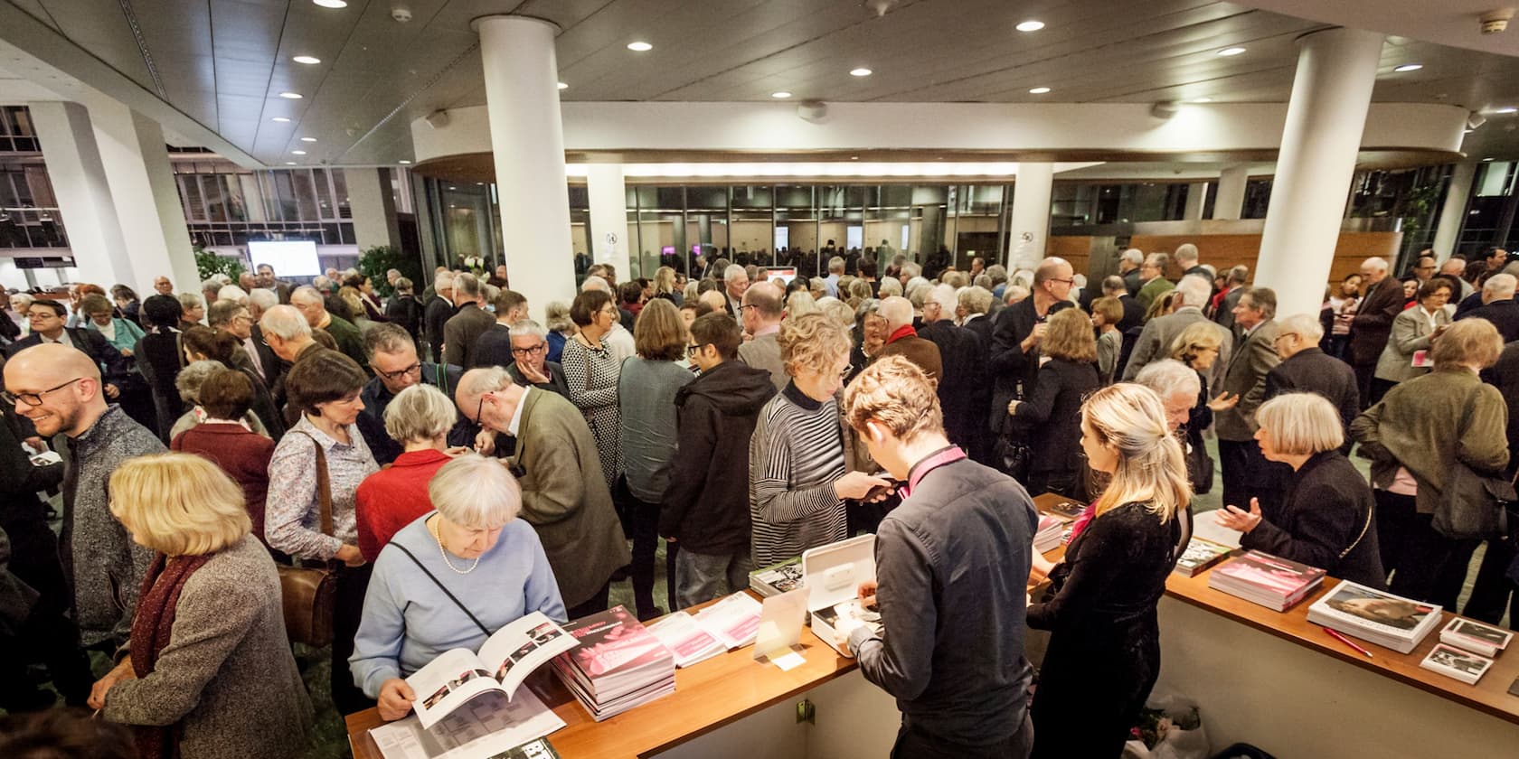 A crowd of people gathered in a well-lit indoor space for an event. Some people are browsing through books on a table.