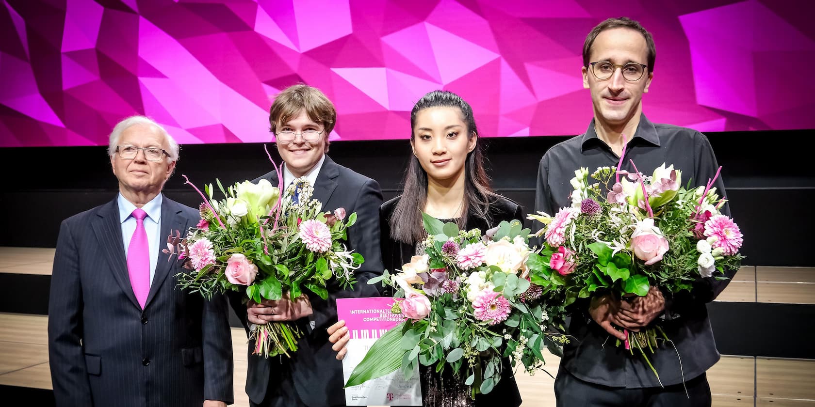 Four people are standing on a stage holding bouquets. The background features a pink geometric wall.