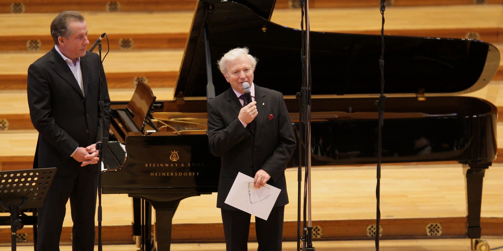 Two men standing on a stage in front of an open grand piano, one holding a microphone and a piece of paper.