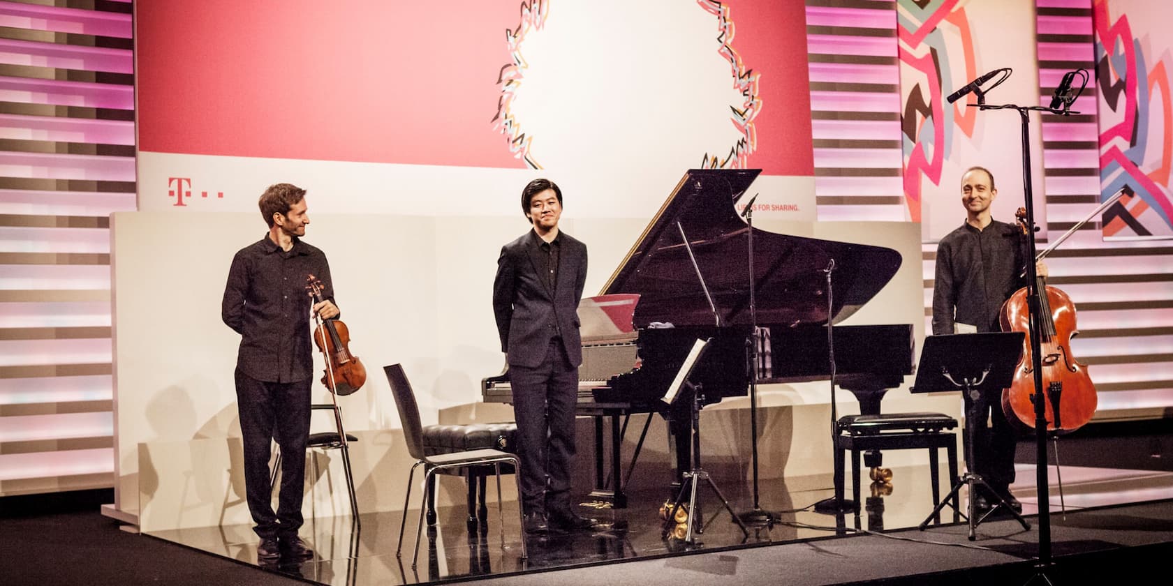 Three musicians on a stage with a grand piano, a violin, and a cello.