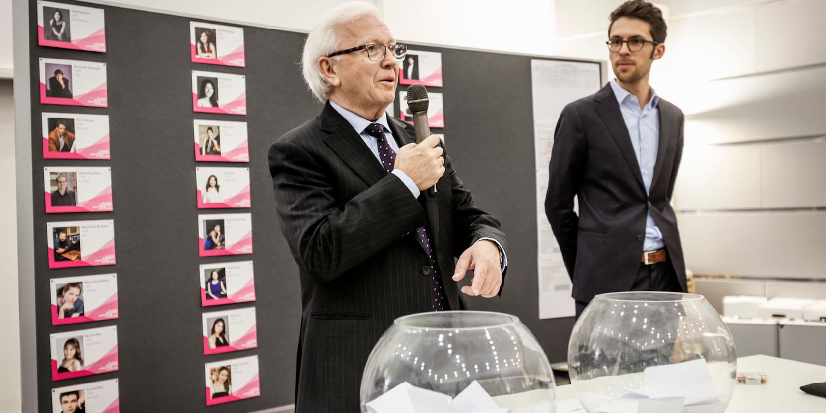 An older man is holding a microphone and speaking, while a younger man stands next to him. In the background, pictures of individuals are displayed on a bulletin board.