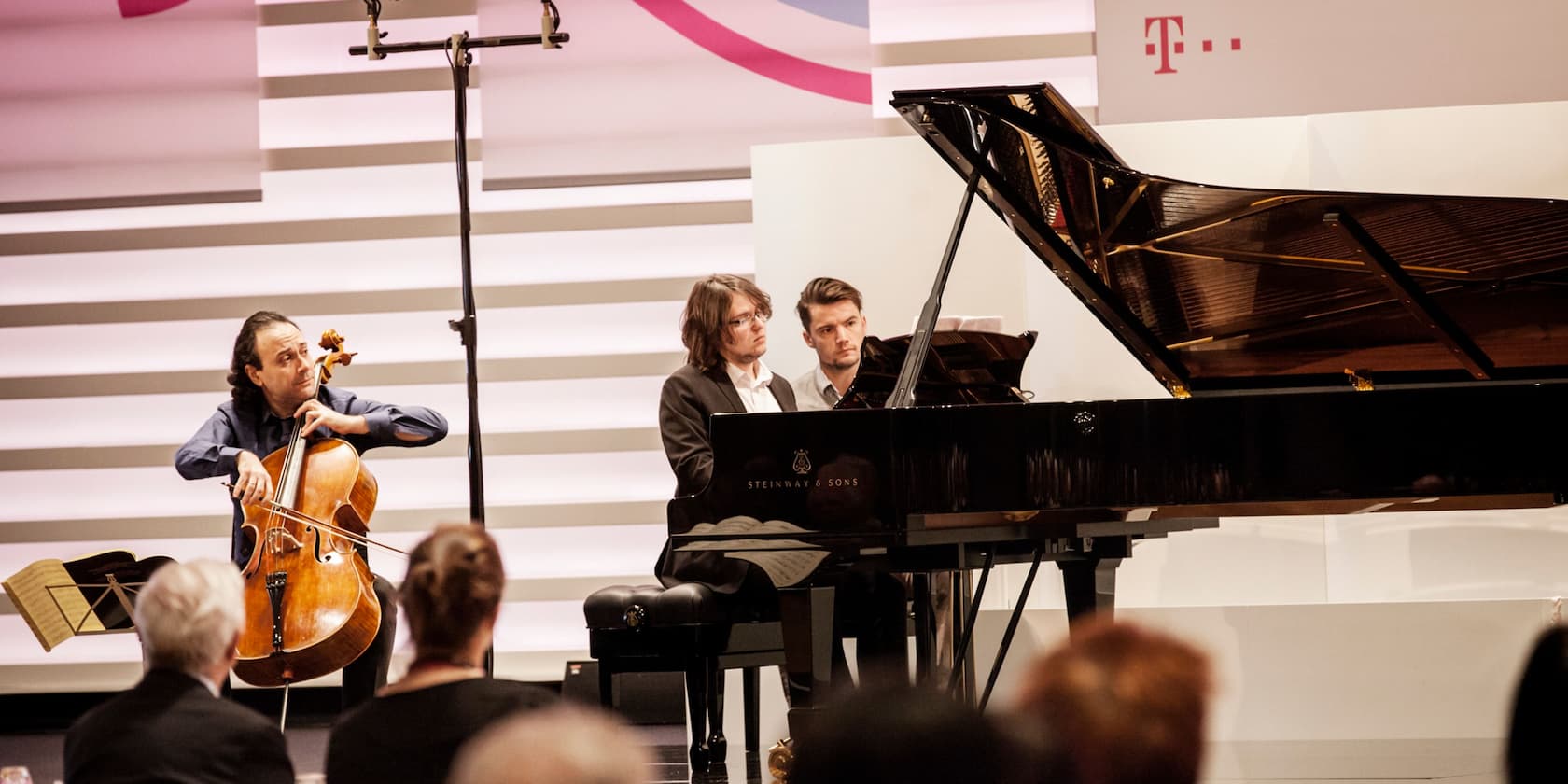 Two musicians playing cello and piano at a concert, with an audience in the foreground.