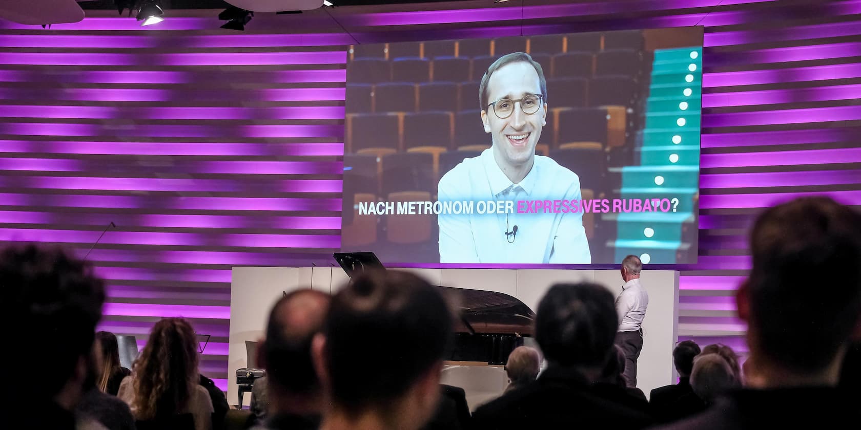 A man speaks on a stage in front of an audience with an image of himself on a large screen in the background. The text on the screen reads: 'NACH METRONOM ODER EXPRESSIVES RUBATO?'