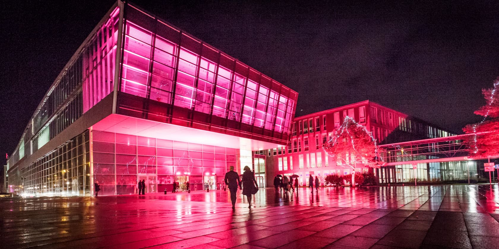 Gebäude bei Nacht mit rosa Beleuchtung, Menschen gehen auf regennassen Boden zu