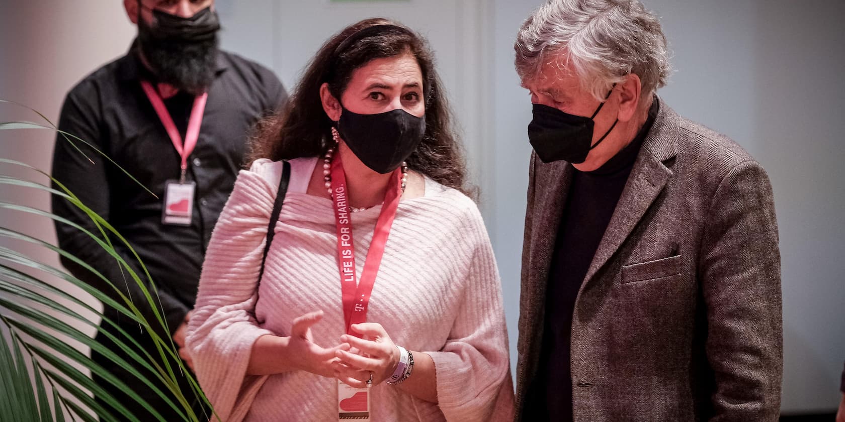 Two people wearing black masks are conversing while another person stands in the background. The woman is wearing a red badge that says 'Life is for sharing'.