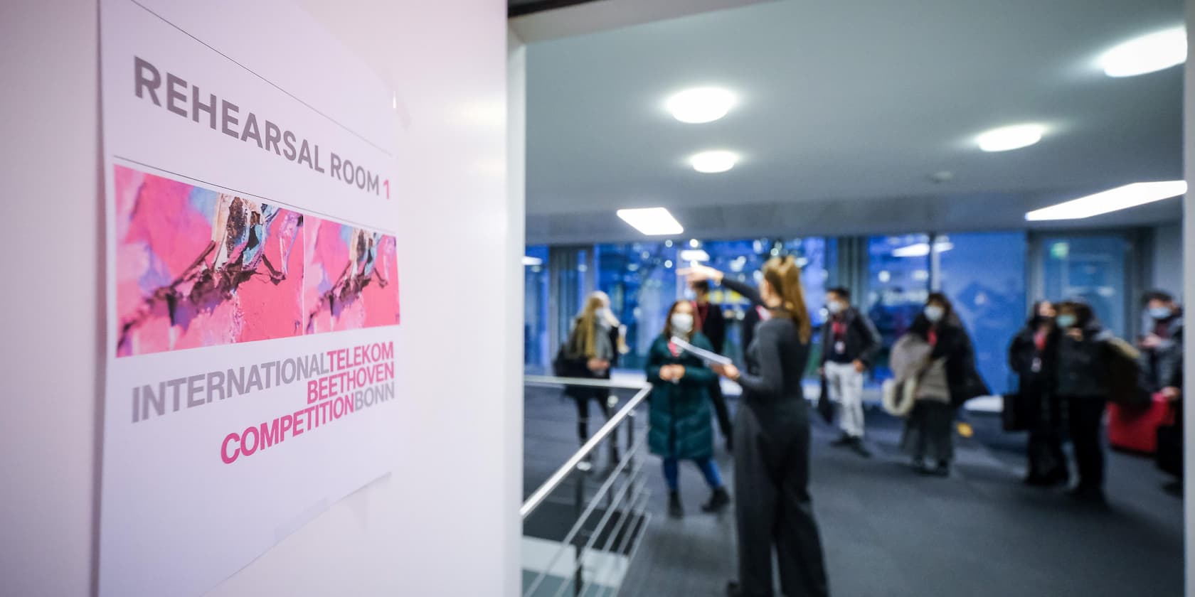 Schild mit der Aufschrift 'Rehearsal Room 1' und 'International Telekom Beethoven Competition Bonn'. Im Hintergrund stehen Menschen in einem Flur.