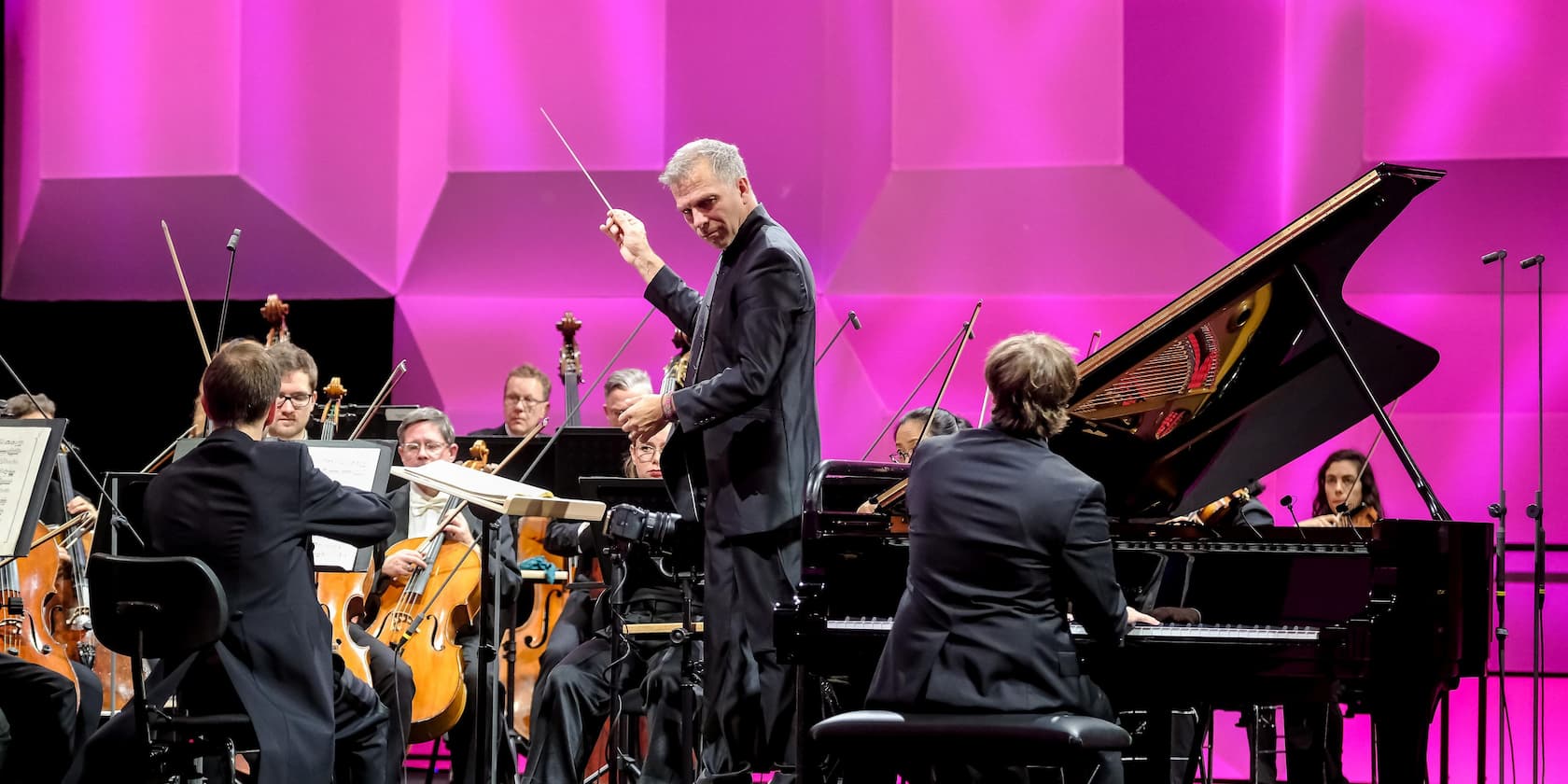 A conductor leads an orchestra playing classical music. In the foreground, a pianist is seen at a grand piano.