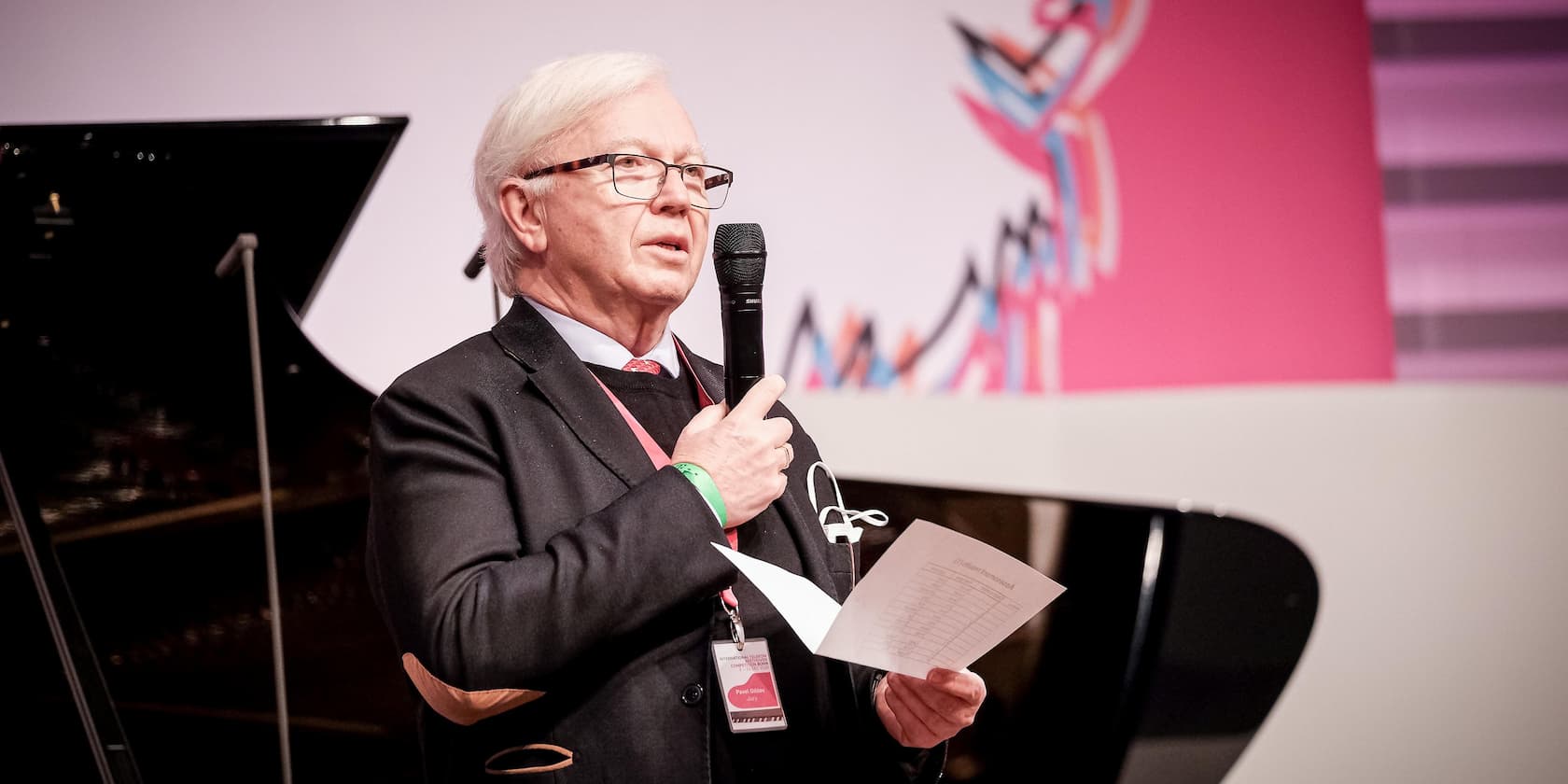 An elderly man holds a microphone and a sheet of paper during a speech.